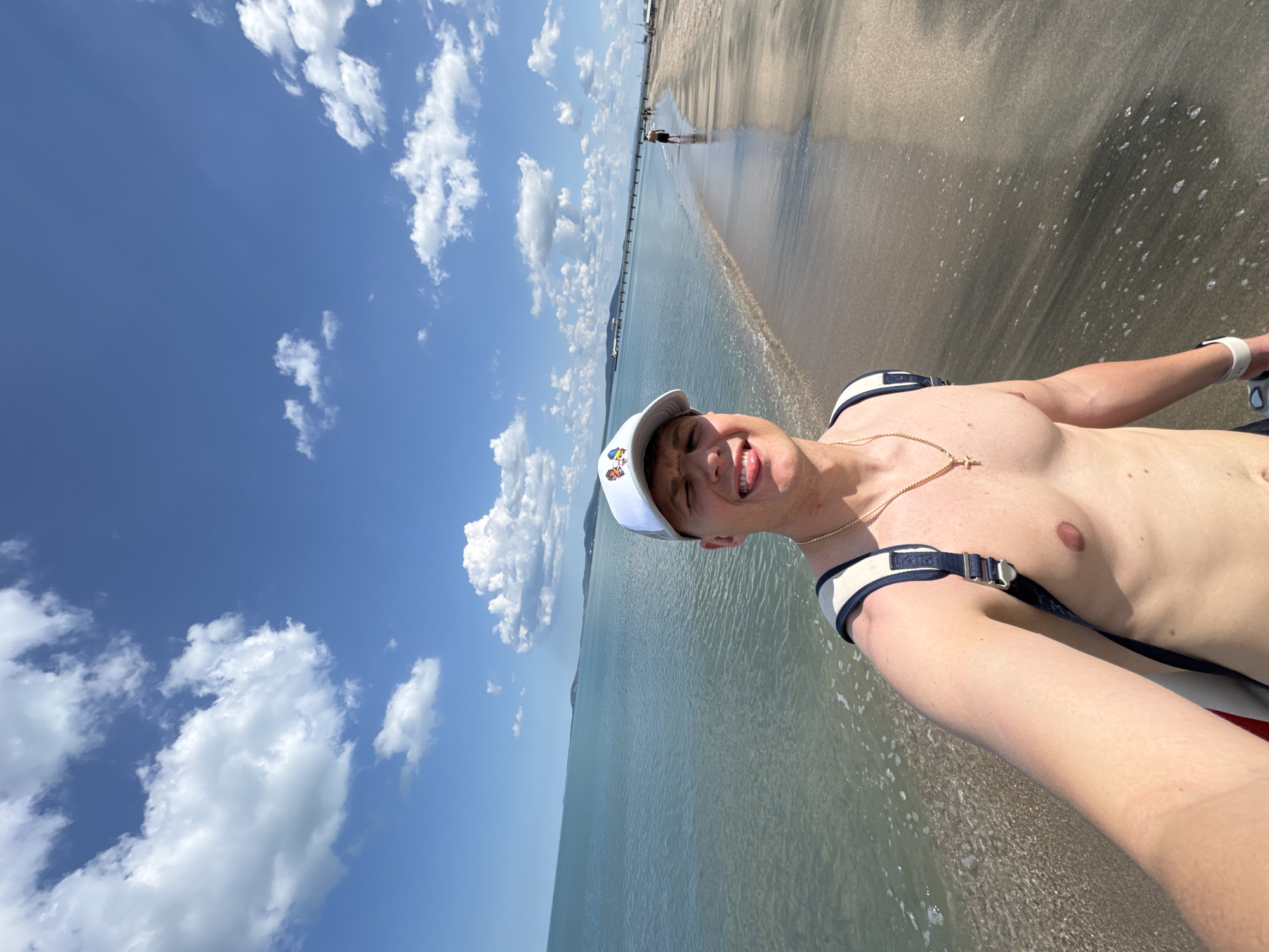 A man is taking a selfie on a beach in Burgas, Bulgaria. He is shirtless, wearing a white cap with cartoon characters, and has a gold chain with a cross pendant. The sky is blue with white clouds. The water meets the beach. In the background, there are other people walking along the shore. A long bridge-like structure is visible extending into the sea in the distance. The beach consists of sand and wet areas where the water has receded.
