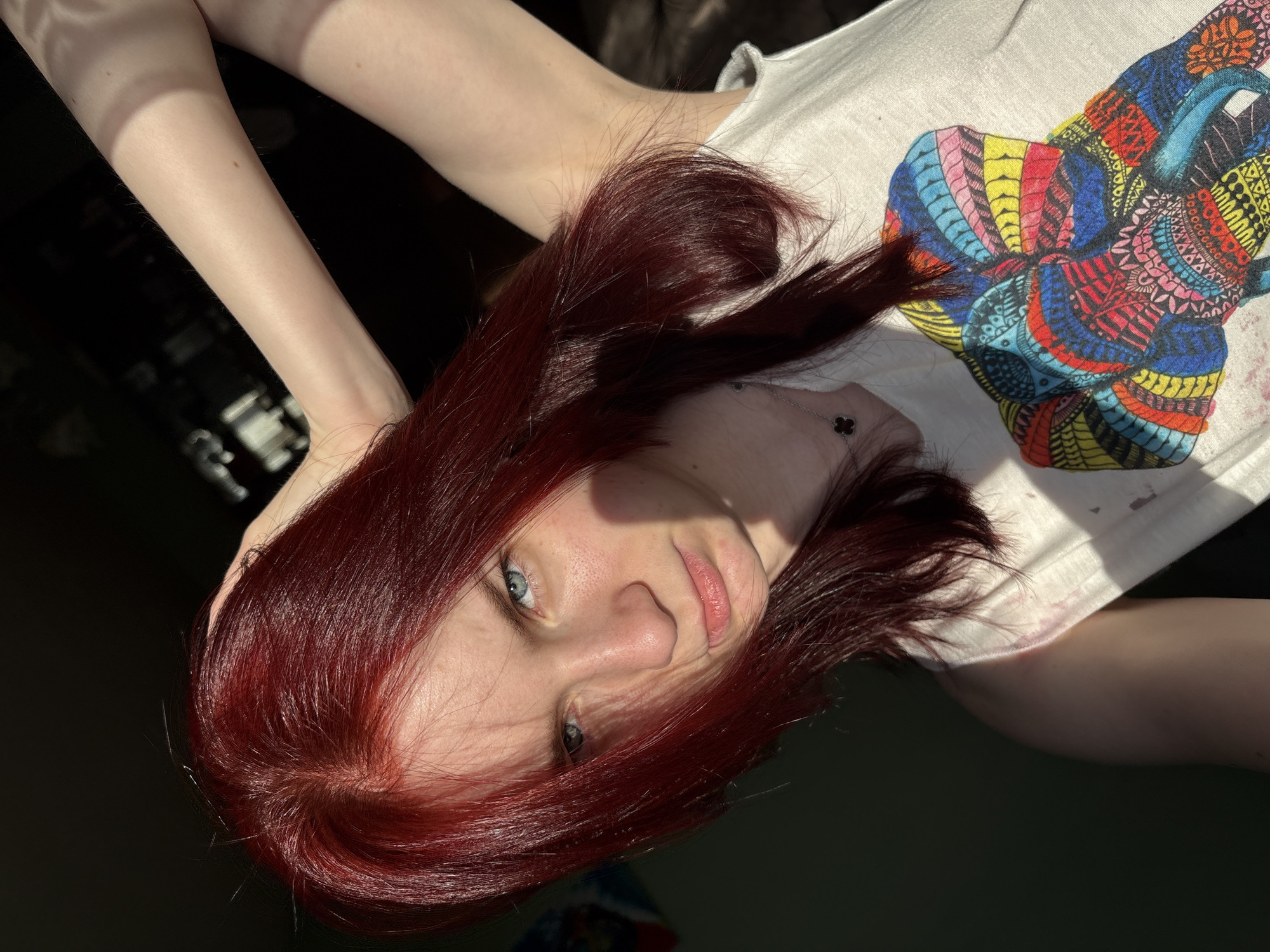 In an indoor setting with dark walls and diffused sunlight casting shadows, a young individual with dark red hair and blue eyes is depicted. The individual is wearing a white t-shirt with a colorful elephant graphic and a delicate necklace. They are positioned in a close-up shot, with one arm raised and hand resting on their head, and the other arm bent, with their hand near their face. The lighting highlights the texture of their hair and skin, creating a dramatic effect. The background is dark and indistinct, with subtle hints of objects and structures.