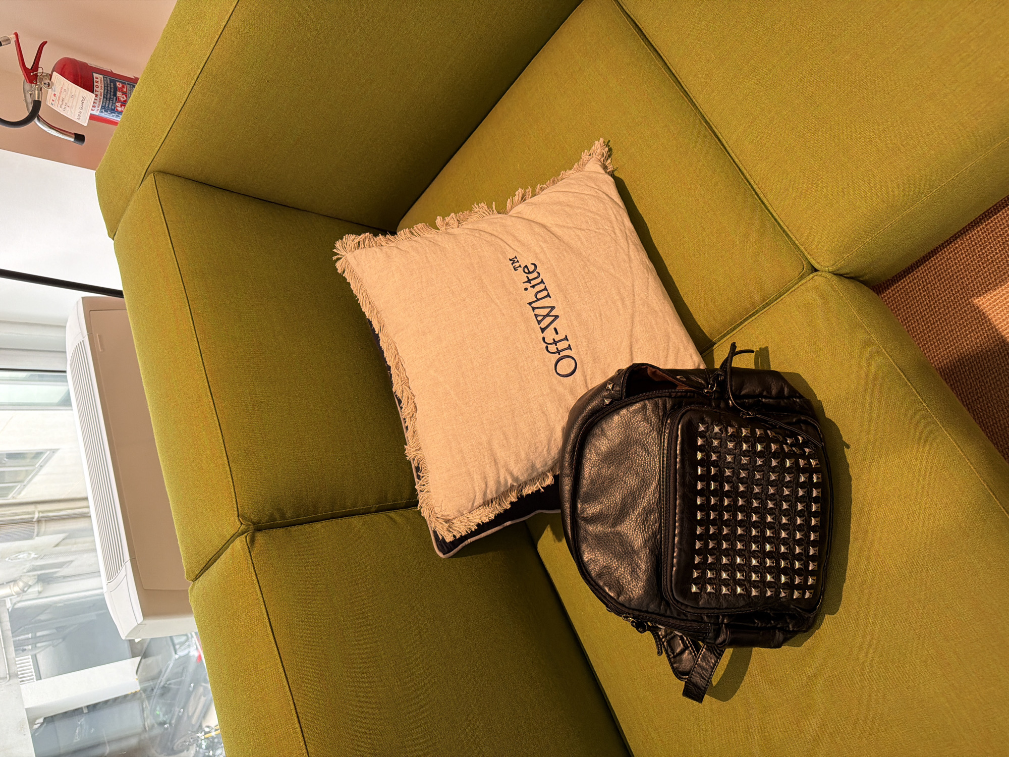 A high-angle, slightly tilted shot captures a lime green modular sofa in an indoor setting, likely a public or commercial space in Milan, Italy. On the sofa, a light beige throw pillow with frayed edges features black embroidered text reading "OFF-WHITE™". Adjacent to the pillow is a black, faux leather backpack or fanny pack, heavily adorned with silver studs on its front pocket.
In the top left, a red fire extinguisher is mounted, its label clearly showing "ESTINTORE" (Italian for fire extinguisher) and "N.U. GUARDIA". Below it, a white electronic unit, possibly an air conditioner, is visible beside a large window offering a glimpse of an exterior building. A textured, brown or tan carpet or rug is partially visible on the far right. The scene is illuminated by natural light, suggesting daytime, with warmer light accentuating the textures on the right side of the sofa and the carpet. No individuals are present, and no specific activity is occurring.