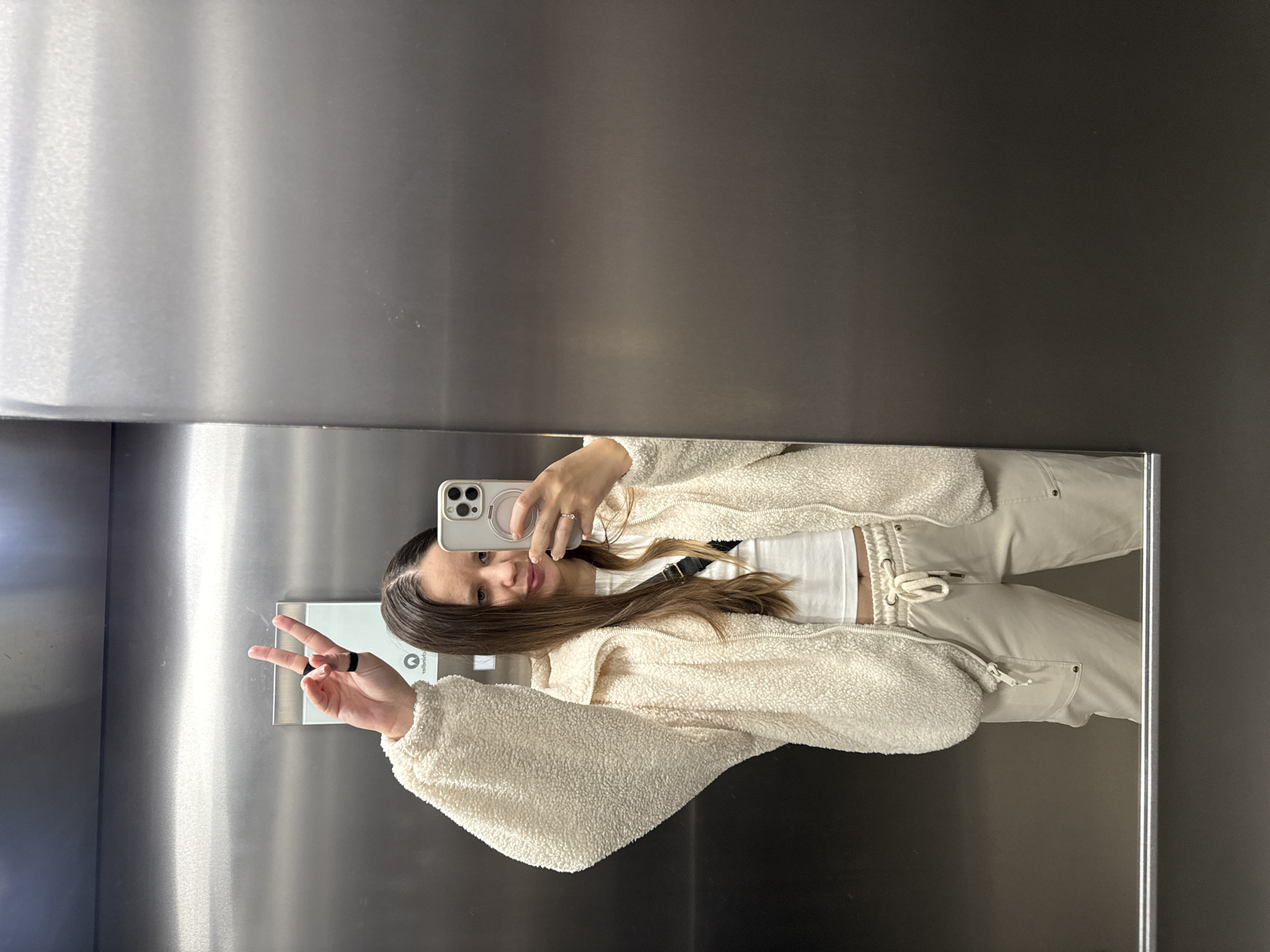 A woman is taking a selfie in an elevator. She's making a peace sign with her left hand and holding a smartphone in her right. She is wearing a white crop top, cream-colored pants with a drawstring waist, and a fleece jacket. Her long brown hair is partially obscuring a black strap across her torso. The elevator walls are metallic. There is no clear indication of the location being in Barcelona, Spain.