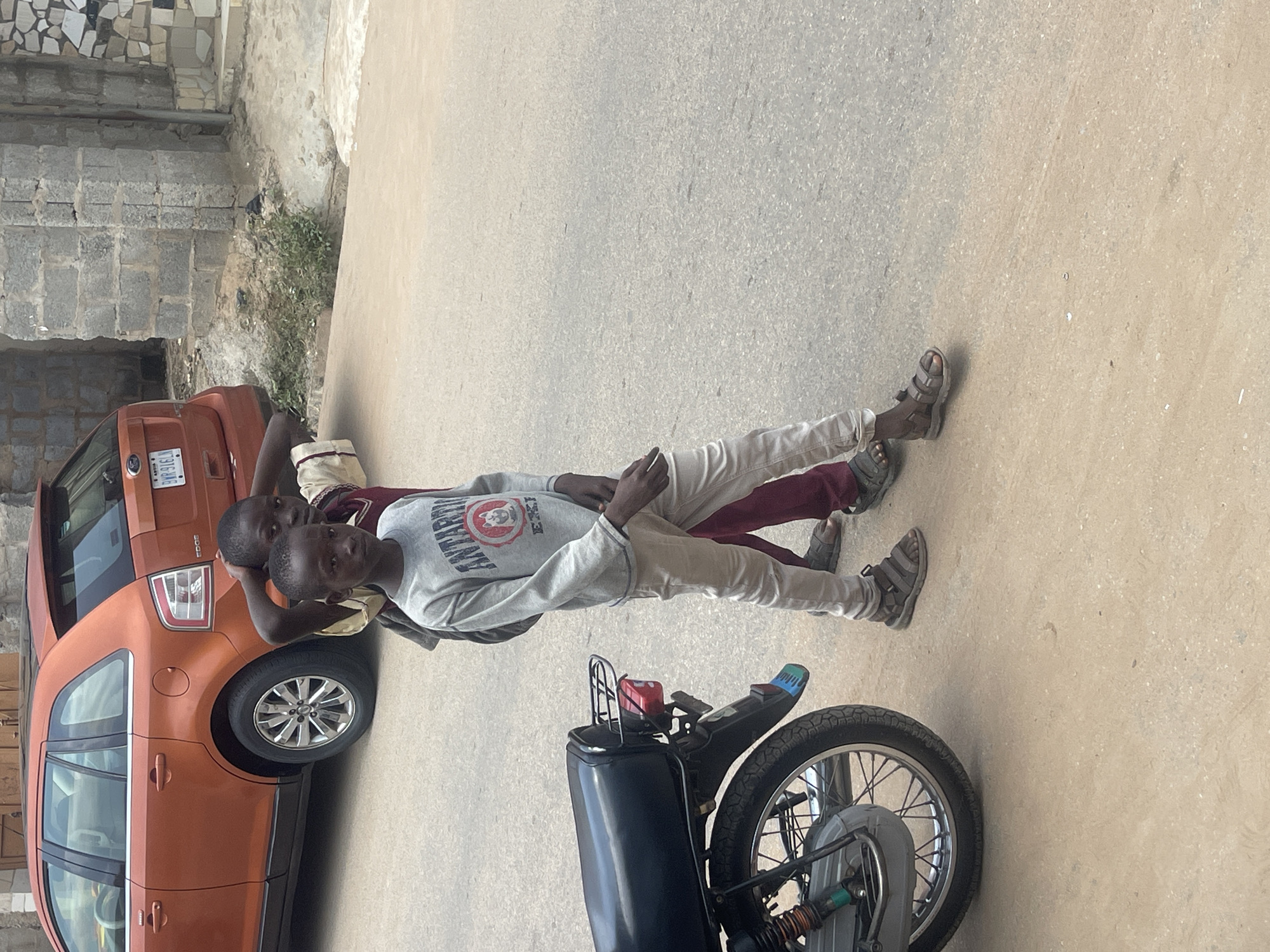 In an outdoor scene on a paved road in Abuja, Nigeria, two young boys are standing. The boy in the foreground, on the left, is wearing a light grey long-sleeved shirt with the word "ANTARCTIC" in dark lettering and a circular graphic, along with light-colored pants and dark sandals. He has his hair cut short and appears to be looking forward. The second boy, slightly behind and to the right, is wearing a maroon shirt and also has short hair. He is touching the back of his head with his left hand and looking towards the camera. To the left of the boys, the rear section of a black motorcycle is visible, showing its wheel, fender, and tail light. In the background, an orange SUV is parked. The environment includes a rough brick wall and a paved surface, suggesting a residential or commercial street.
