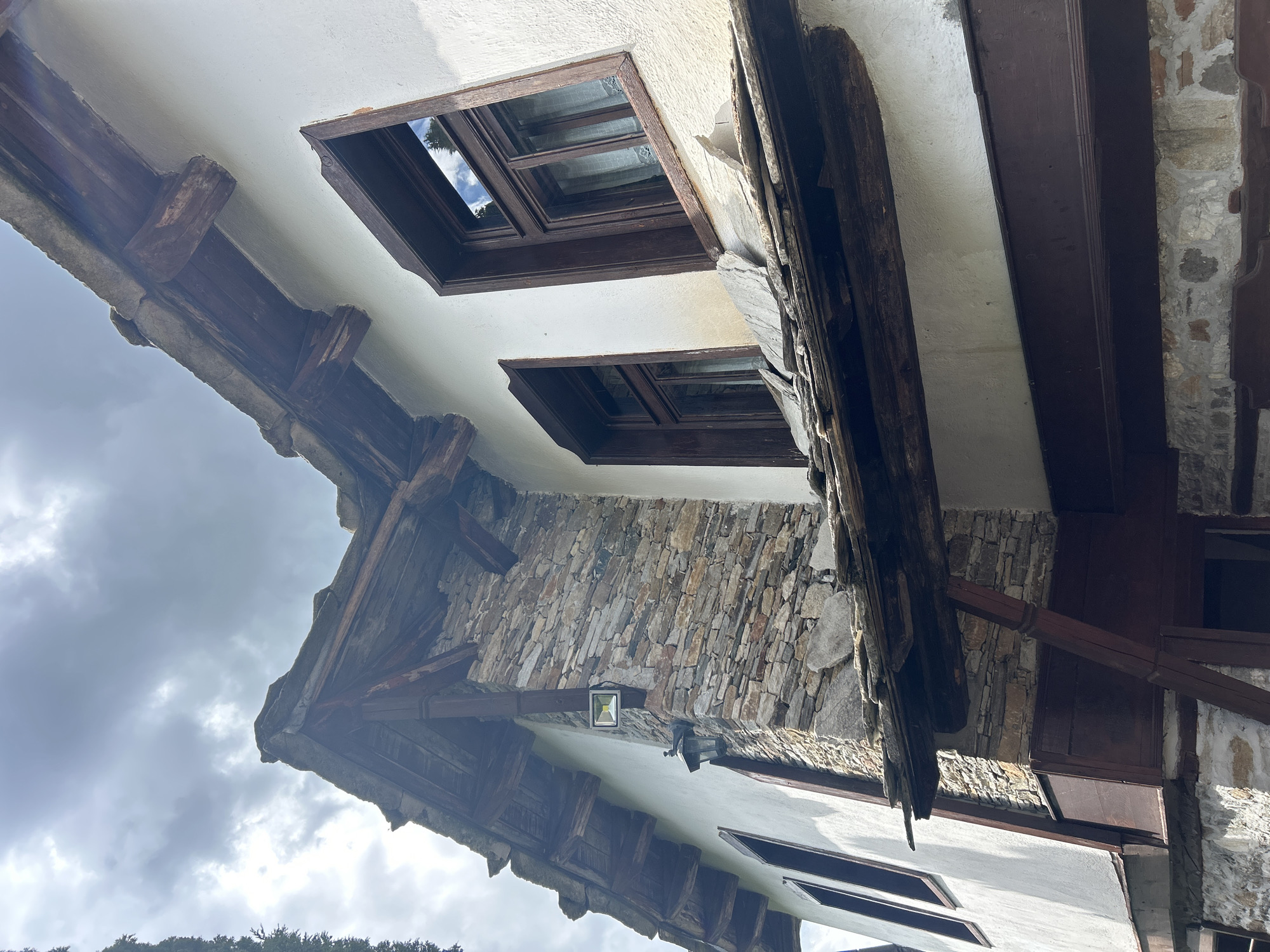 The image depicts the exterior architecture of a building in the village of Dolen, Bulgaria, known for its well-preserved Bulgarian National Revival architecture. The building features a combination of white stucco walls and stone masonry. Distinct elements include dark wood-framed windows with reflections of the sky, exposed wooden beams, and a stone gable end. The lighting fixtures, including a square spotlight and a traditional lantern, are visible. The sky is overcast, with visible clouds.