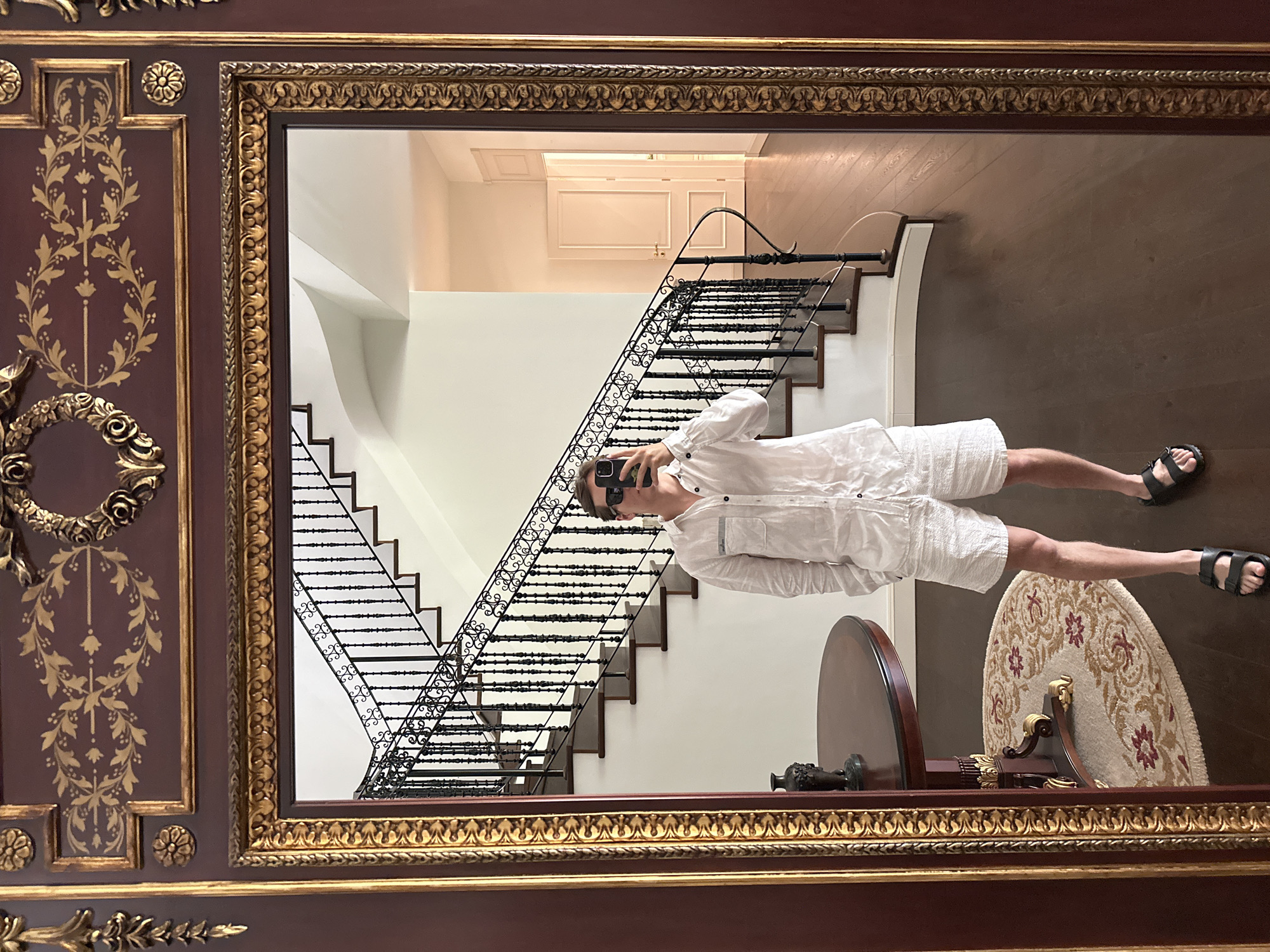 A person is taking a mirror selfie in a grand foyer with a large, ornate mirror. The person is wearing a white linen shirt and white textured shorts with black sandals. They are holding a smartphone up to take the photo. In the mirror's reflection, a large staircase with a black ornate railing is visible. To the left of the staircase, a round, dark wood table with a decorative vase sits on a circular rug. In the background of the reflection, a hallway with several doors is visible. The architecture suggests a luxurious residence.