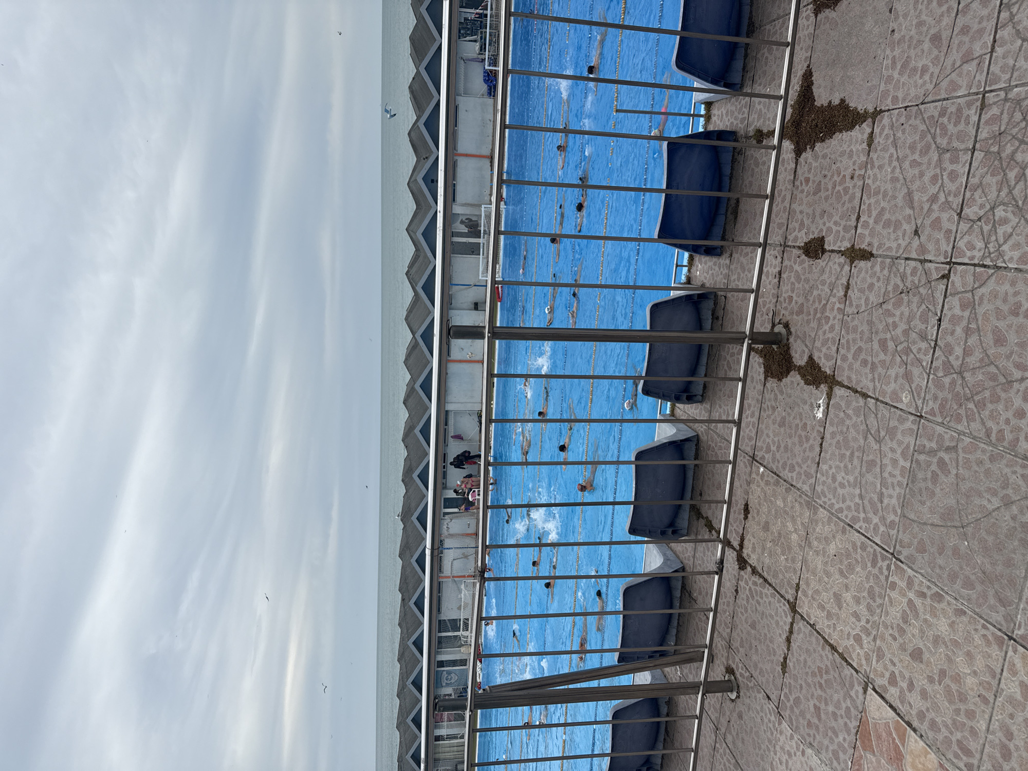 From an elevated perspective, the photograph captures the Primorski swimming pool complex in Varna, Bulgaria, with a partial view of the Black Sea coast and overcast sky. The pool area is occupied by swimmers practicing laps in multiple lanes, which are visible through a metal railing and a row of blue, stadium-style seats. The building housing the pool, identifiable by its sawtooth roofline, is adjacent to the swimming area. The foreground features tiled paving, marred by accumulated debris, indicating a state of disrepair or incomplete maintenance. A variety of individuals are visible in the pool and around it, suggesting activity during the day.