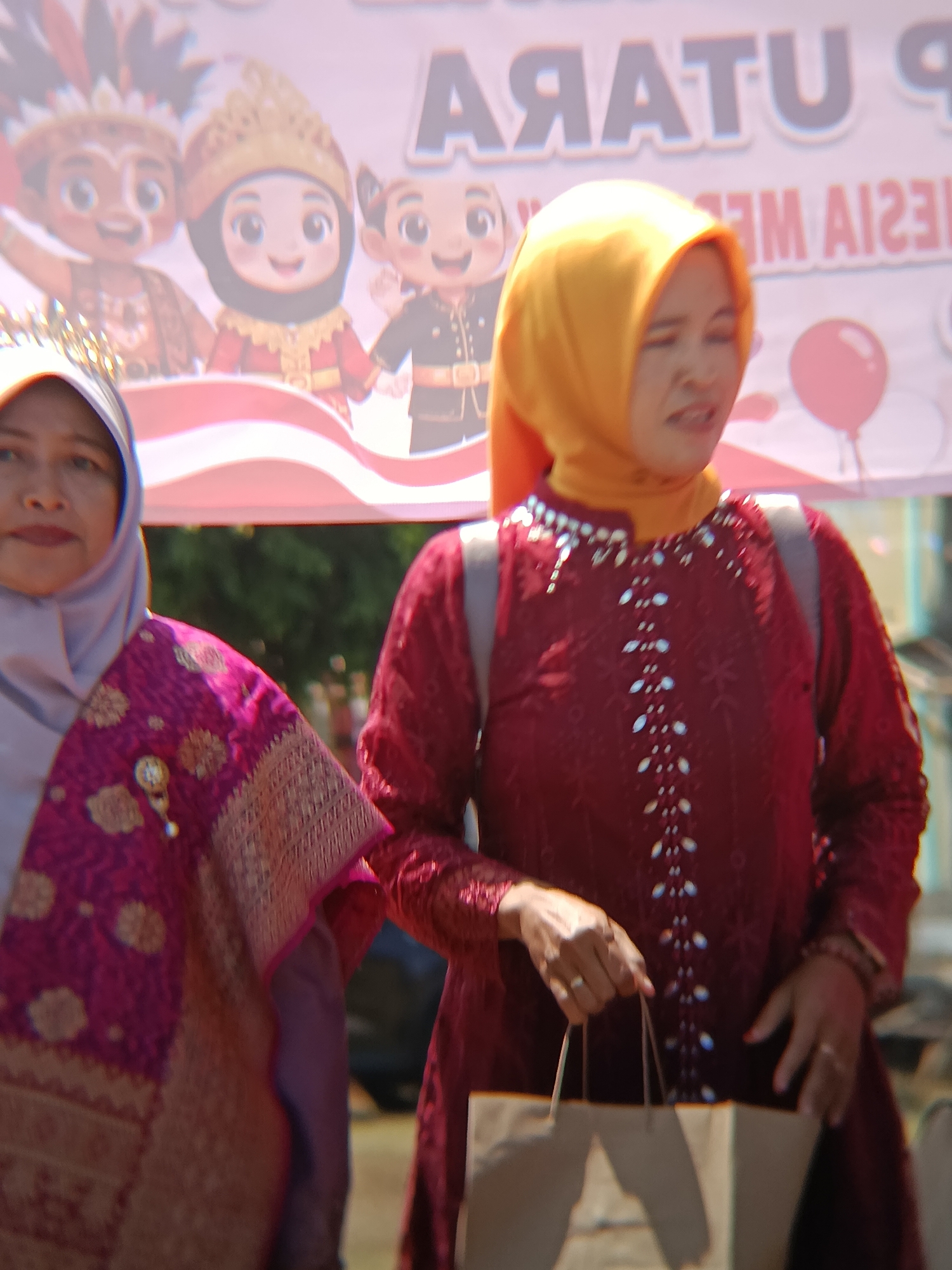 In an outdoor setting in Desa Kebon Manis, Kecamatan Cilacap Utara, Indonesia, two women are prominently featured in the foreground. The woman on the left, wearing a light purple hijab and a patterned fuchsia garment adorned with a brooch, is partially visible, her face turned towards the viewer. To her right, a woman wearing a bright yellow hijab and a dark red, long-sleeved garment with intricate beading stands looking off to the side with a strained expression. She is holding a light brown paper bag with both hands. In the background, a blurred banner displays text in what appears to be Indonesian, featuring cartoon characters in traditional attire. Red and white stripes are visible on the banner, suggesting a festive or nationalistic theme. The environment appears to be a public space, possibly a street or a gathering area, with hints of greenery and other figures blurred in the distance.