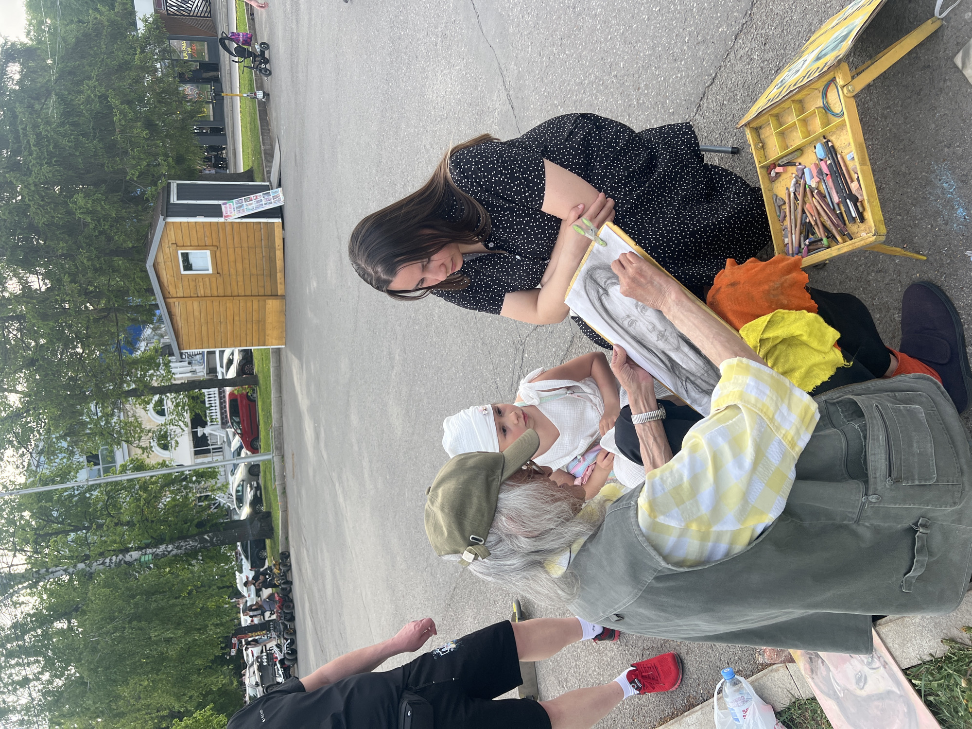 The image shows an outdoor scene in an urban area, possibly Ufa, Russia. A young woman in a black and white polka dot dress is observing an artist drawing a portrait of a young girl wearing a white headscarf. The artist, an older person with gray hair and an olive green cap, is sitting on the ground and holding a drawing board with a charcoal portrait in progress. Next to the artist is a yellow open box containing drawing materials. An additional person is partially visible in the foreground wearing black shorts and red shoes. The background includes parked cars, a small wooden building, and trees. The ground is paved, suggesting a street or sidewalk setting.