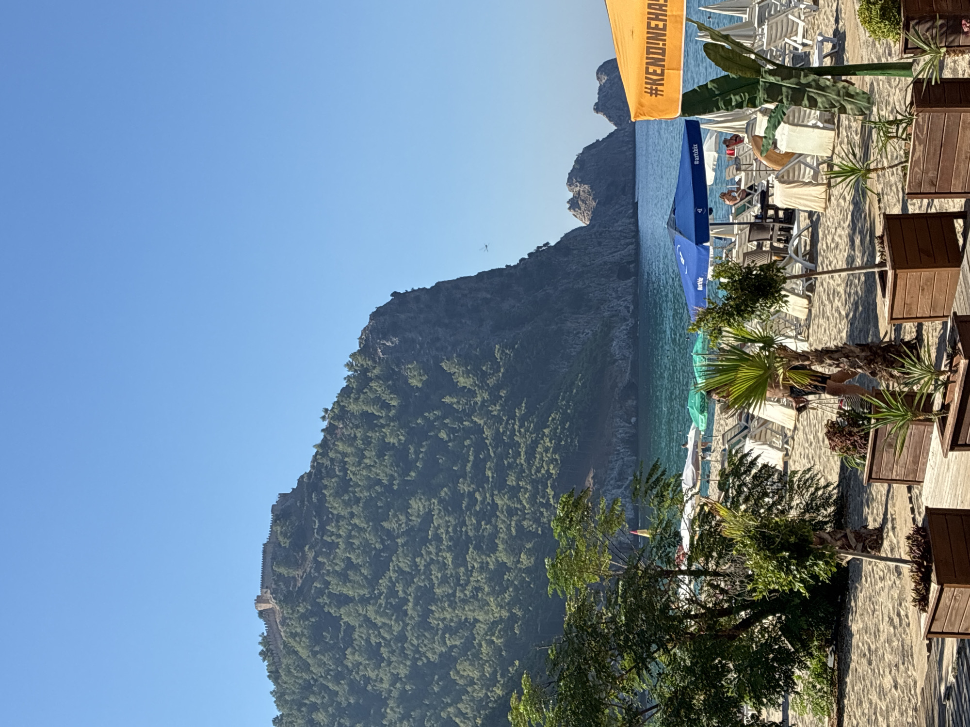 A scenic view of a beach in Alanya, Turkey, shows a sandy shore dotted with lounge chairs and umbrellas. In the background, a lush, tree-covered mountain rises dramatically from the turquoise sea, crowned by a historic castle ruin. The clear blue sky suggests a sunny day. The foreground features wooden planters with various plants, and a portion of an orange umbrella with "#KENDI!NEHAS" written on it. Several people are visible on the beach, relaxing on lounge chairs or walking. The sea is calm, with small waves lapping the shore.