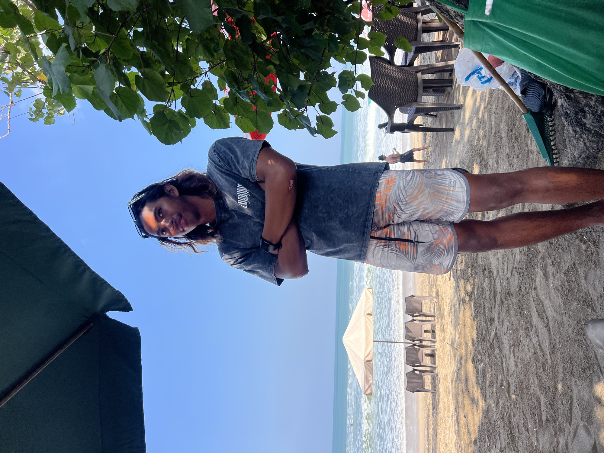 A young man with long, brown hair and sunglasses perched on his head stands on a sandy beach in Kuta, Indonesia, with his arms crossed. He is wearing a dark gray t-shirt with a white graphic and patterned shorts. Behind him, the ocean is visible with gentle waves and a clear blue sky above. Several empty beach chairs are arranged on the sand, some under a light-colored umbrella. To the right, lush green leaves from a tree overhang part of the scene, and further back, wicker chairs and a rake are visible. A few other figures are faintly visible in the distance on the beach, near the water.