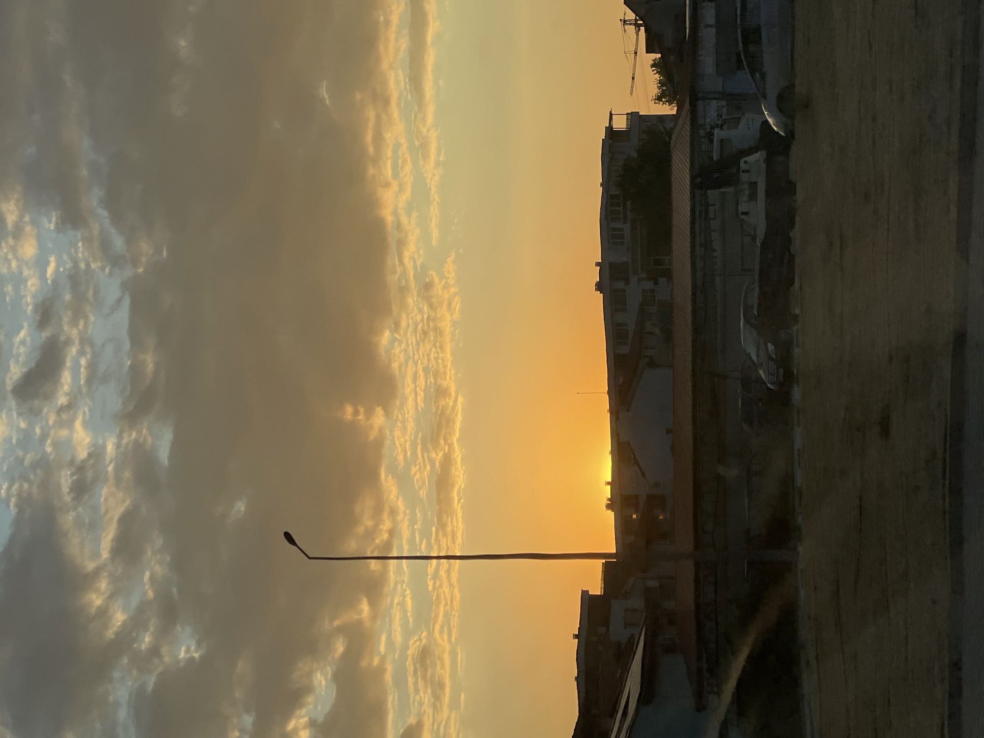 The image captures a sunset scene in Asenovgrad, Bulgaria. The sky is filled with a gradient of colors, from deep orange at the horizon where the sun is setting, to lighter yellow and then dusky gray clouds towards the top. A tall, slender lamppost stands prominently on the left side of the frame, silhouetted against the bright sky. In the mid-ground, several buildings are visible, appearing to be apartment blocks and other structures characteristic of an urban environment. The lower part of the image shows a street or an open area with several parked vehicles, including cars and a truck. The overall atmosphere is serene, with the fading light casting a warm glow over the cityscape.