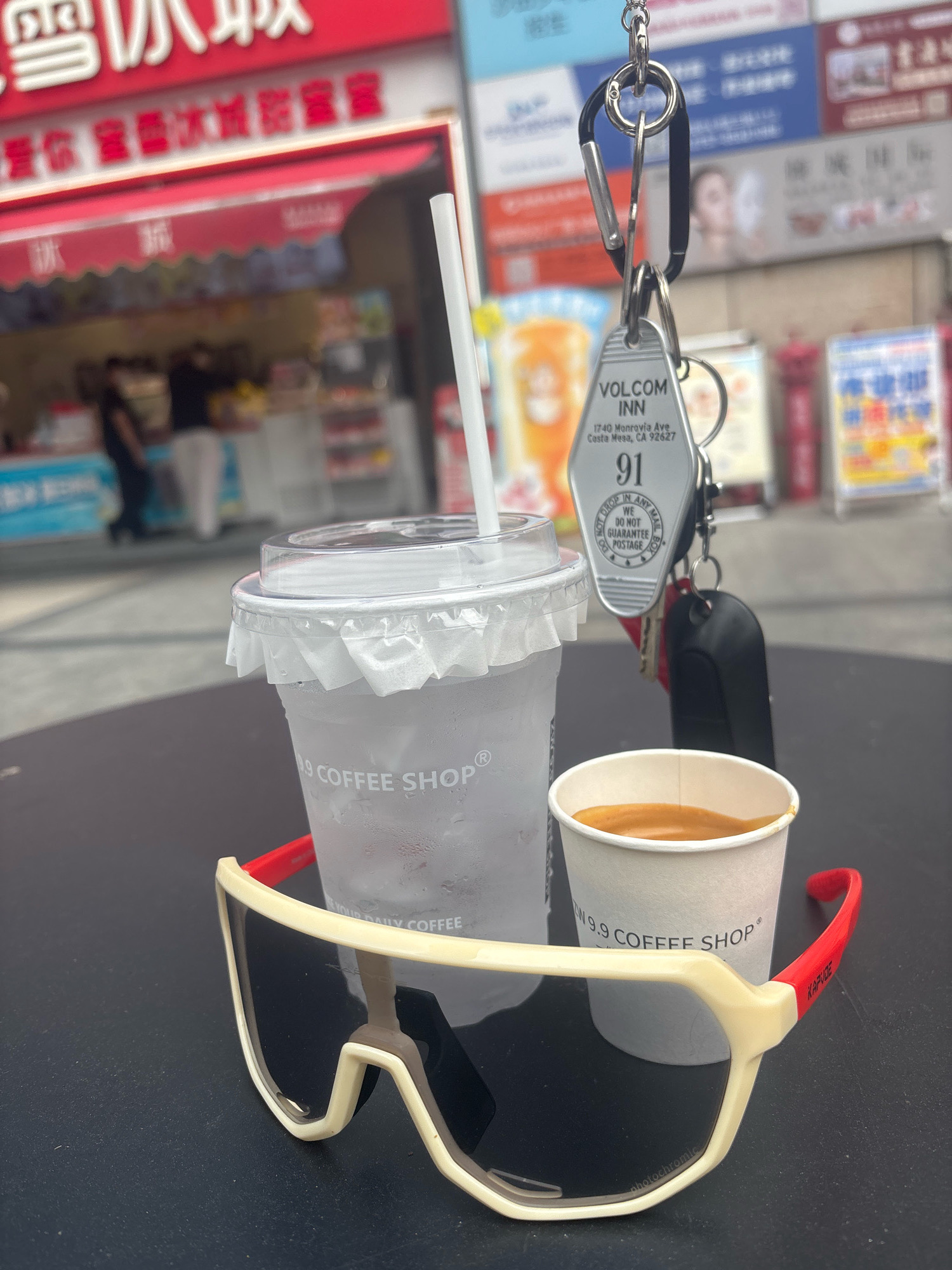 In the city of Nanhai District, China, the image captures a close-up view of items placed on a dark surface, likely a table. In the foreground, a pair of sunglasses with thick, off-white frames and red accents rests on the surface. Behind the sunglasses, a white paper cup, partially filled with a dark liquid, is visible. To the left, a clear plastic cup, filled with ice and a beverage, has condensation on its exterior and a white straw inserted. A white paper collar is wrapped around the top of the plastic cup. Hanging from a carabiner clipped to a keyring, a silver hotel key tag for "VOLCOM INN" with an address in Costa Mesa, CA is prominently displayed. Two car keys are also attached to the keyring. In the background, a storefront with red signage featuring Chinese characters is blurred. The signage appears to be for a food or beverage establishment. Further in the background, out-of-focus figures of people are present, suggesting a public outdoor setting. The overall lighting indicates daylight.
