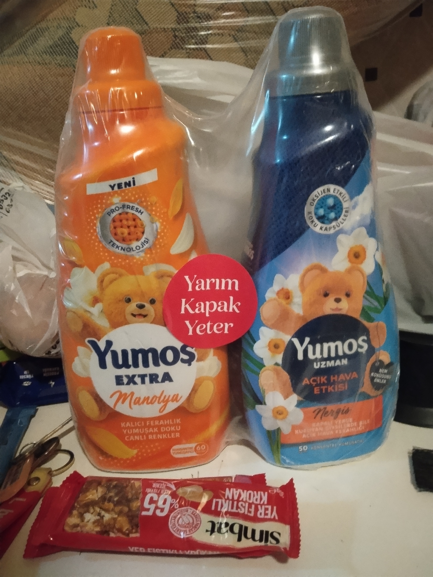 The image shows two bottles of Yumoş fabric softener, one orange (Manolya) and one blue (Nergis), a red sticker with the text "Yarım Kapak Yeter," and a red "Simbat Yer Fıstıklı Krokan" candy bar, all on a white surface. The items are likely indoors. The presence of the Yumoş products, popular in Turkey, and the Simbat candy bar, also a Turkish product, suggest the location is in Turkey. Given the context of the prompt, the location is likely Sarıyer, Turkey, though this cannot be confirmed visually.