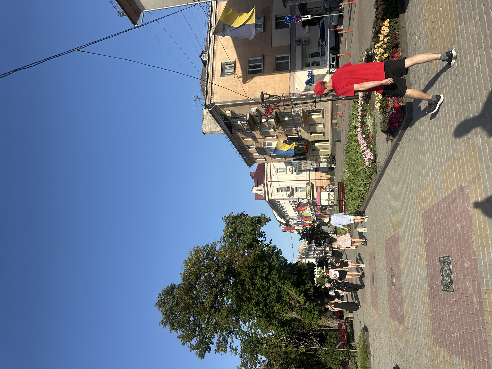 The image depicts a sunny day on a pedestrian walkway in Ternopil, Ukraine. A group of people are walking on the colorful brick pathway, some with dogs. Buildings line the street, adorned with flags. One man in a red shirt and black shorts walks away from the camera. The environment is urban, with trees, flowers, and streetlights visible. The sky is a clear blue.