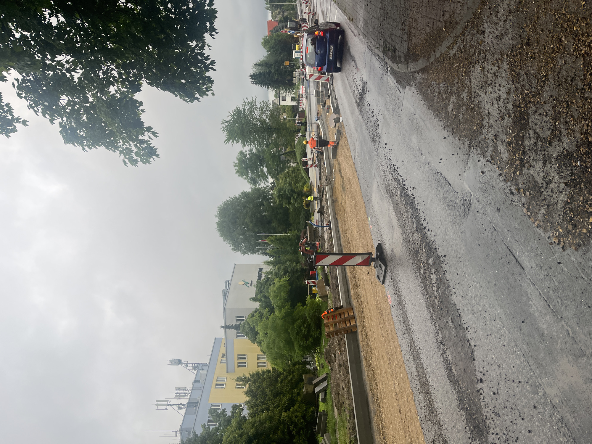 The image depicts a street construction scene in a city. A blue car is driving on a wet asphalt road. The road is partially under construction, with gravel and some paving stones visible. There are construction barriers, including red and white striped traffic cones and signs, placed along the road. Several construction workers in high-visibility vests and helmets are present, some appearing to be working on the road or sidewalk area. In the background, there is a building with a yellow and gray facade, and lush green trees line the street. The sky is overcast, suggesting a cloudy or potentially rainy day.