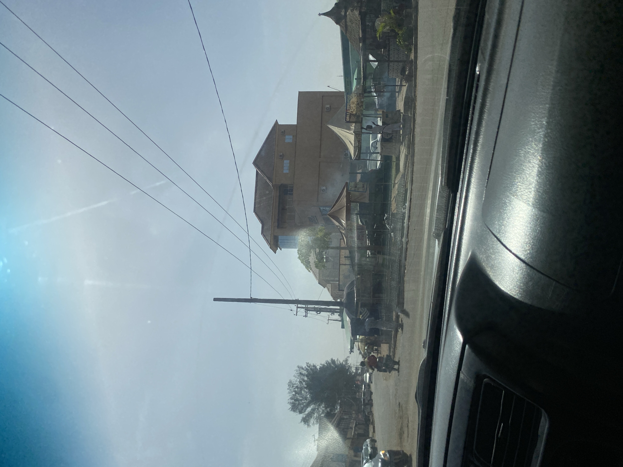 The image captures a vibrant street scene in Barden Yana, Nigeria, viewed from inside a vehicle on a bright, sunny day. The dark interior of the car's dashboard and windshield frame is visible along the right edge of the frame.
The central feature is a multi-story building with light-colored walls and a reddish-brown pitched roof, featuring several visible air conditioning units. In front of this building, along the street, several white or light-colored awnings and tents are set up. Under one of these awnings, smoke is visibly rising, indicating an outdoor cooking or grilling operation. A faint sign on one of the awnings reads "GRILL & SIPS."
Numerous power lines traverse the clear, bright sky, originating from a tall utility pole positioned on the left side of the frame. Below the power lines, a large tree with dense, dark foliage stands by the roadside.
The street is bustling with activity, showing several people walking or standing. A person on a motorcycle is visible further down the road, and other vehicles can be seen in the distance. The scene depicts everyday life in a local commercial or residential area, with probable outdoor food vendors or market stalls operating under clear skies.