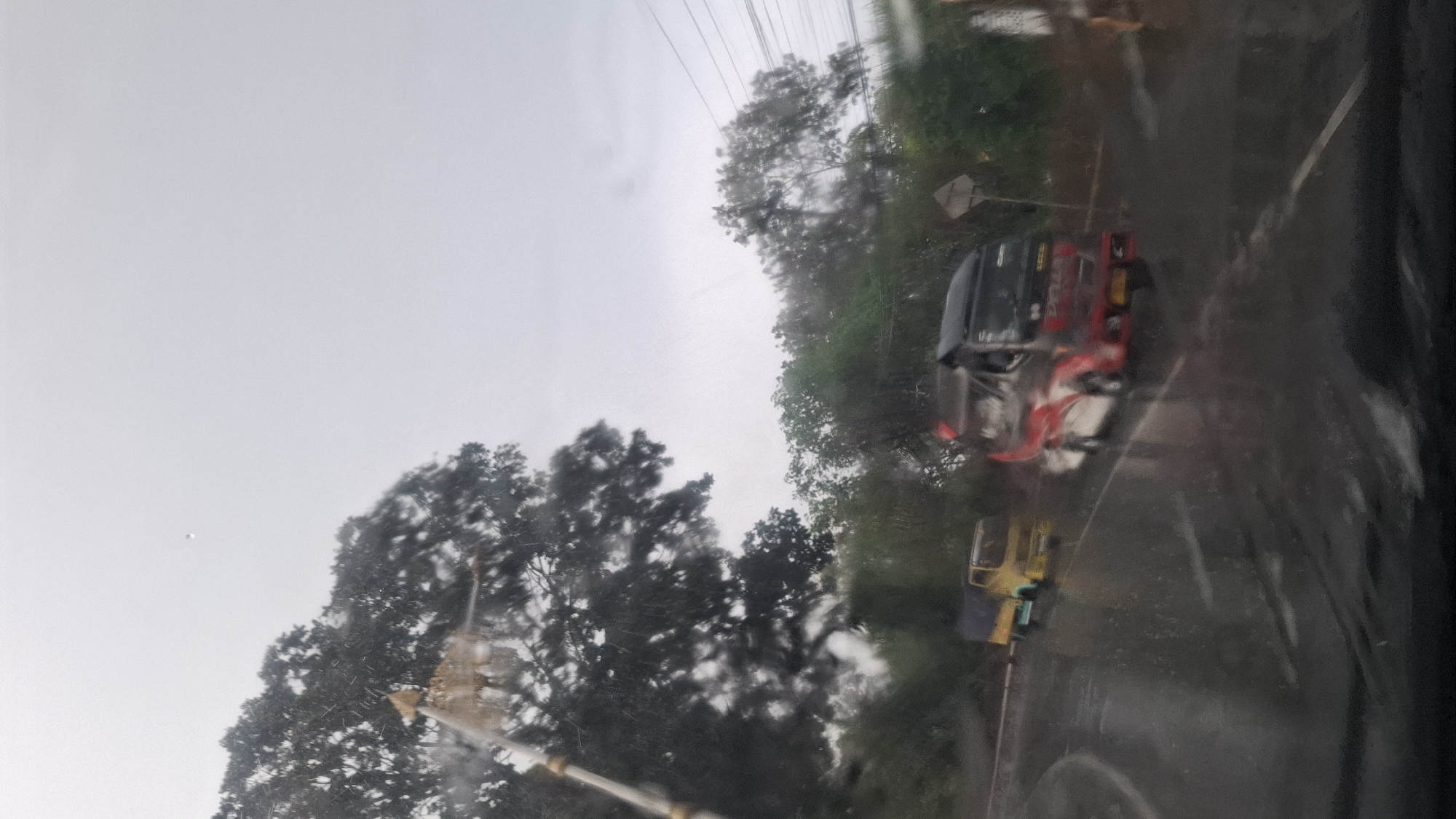 The image depicts a rainy day scene on a road, possibly in Cilaku, Indonesia. A large tree with dark foliage is visible on the left side of the frame, with its branches extending towards a cloudy sky. In the foreground, a wet road surface is apparent, with streaks of water suggesting heavy rainfall.
Two vehicles are present on the road. On the left, a small yellow truck with a blue tarp on its cargo bed is driving away from the camera. To its right and slightly further down the road, a larger truck with a black and red striped design is moving towards the camera. The truck has "SUMBER" visible on its side. The environment appears to be a somewhat rural or suburban area, with lush green trees and vegetation lining the sides of the road. Power lines are also visible in the upper right portion of the image. The overall impression is one of a gray, overcast day with poor visibility due to the rain.