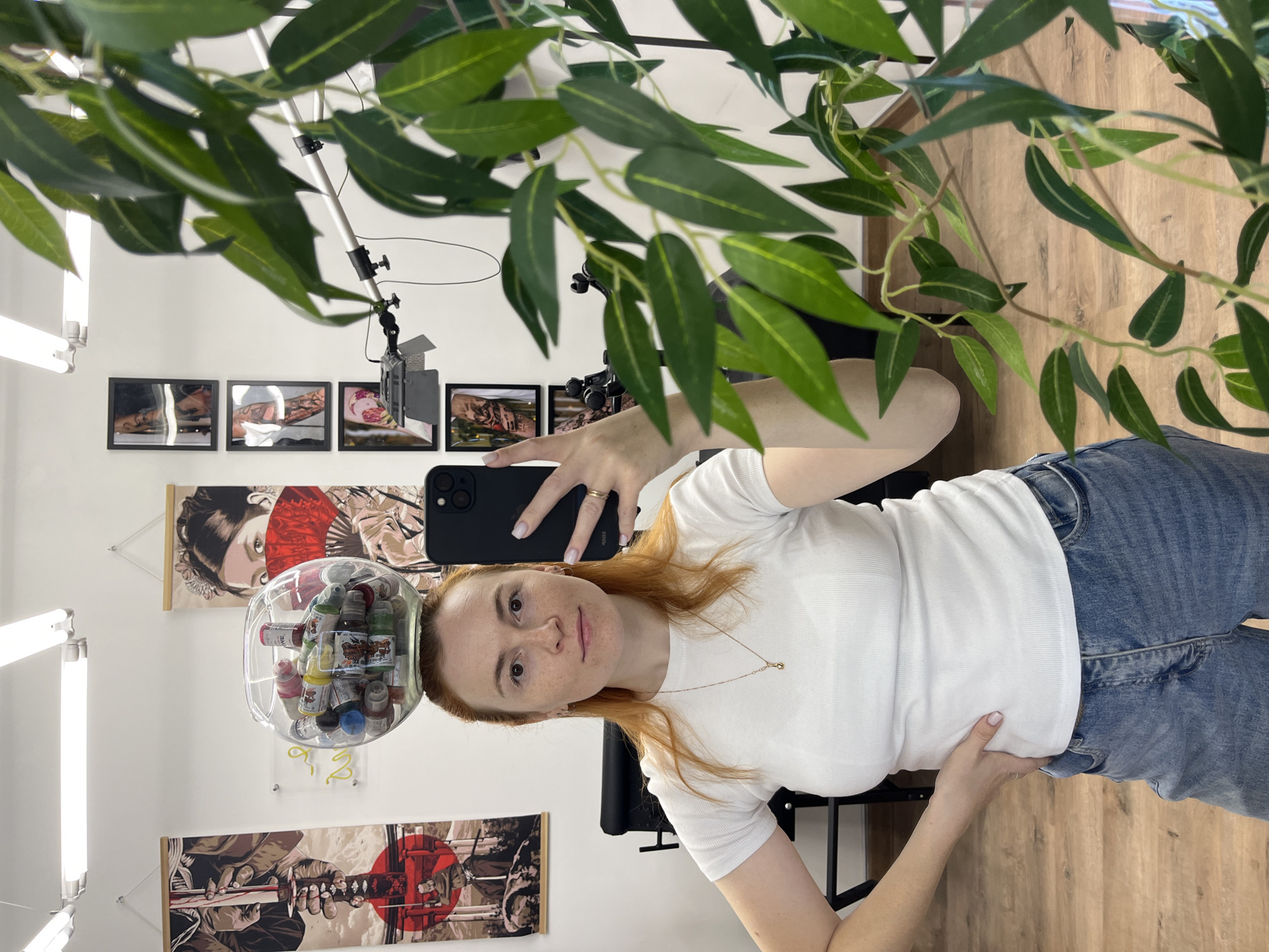 A young woman with red hair stands in front of a mirror, taking a selfie. She is wearing a white t-shirt and blue jeans. A glass bowl filled with small bottles of colorful liquid is balanced on her head. The background appears to be a tattoo studio, with artwork on the walls and a tattoo chair visible. A plant with green leaves partially obscures the right side of the image. The lighting is bright, coming from overhead fixtures. The woman is looking directly at the camera with a neutral expression. The floor is made of wood. There is no indication of the city of Chaikovsky, Russia.