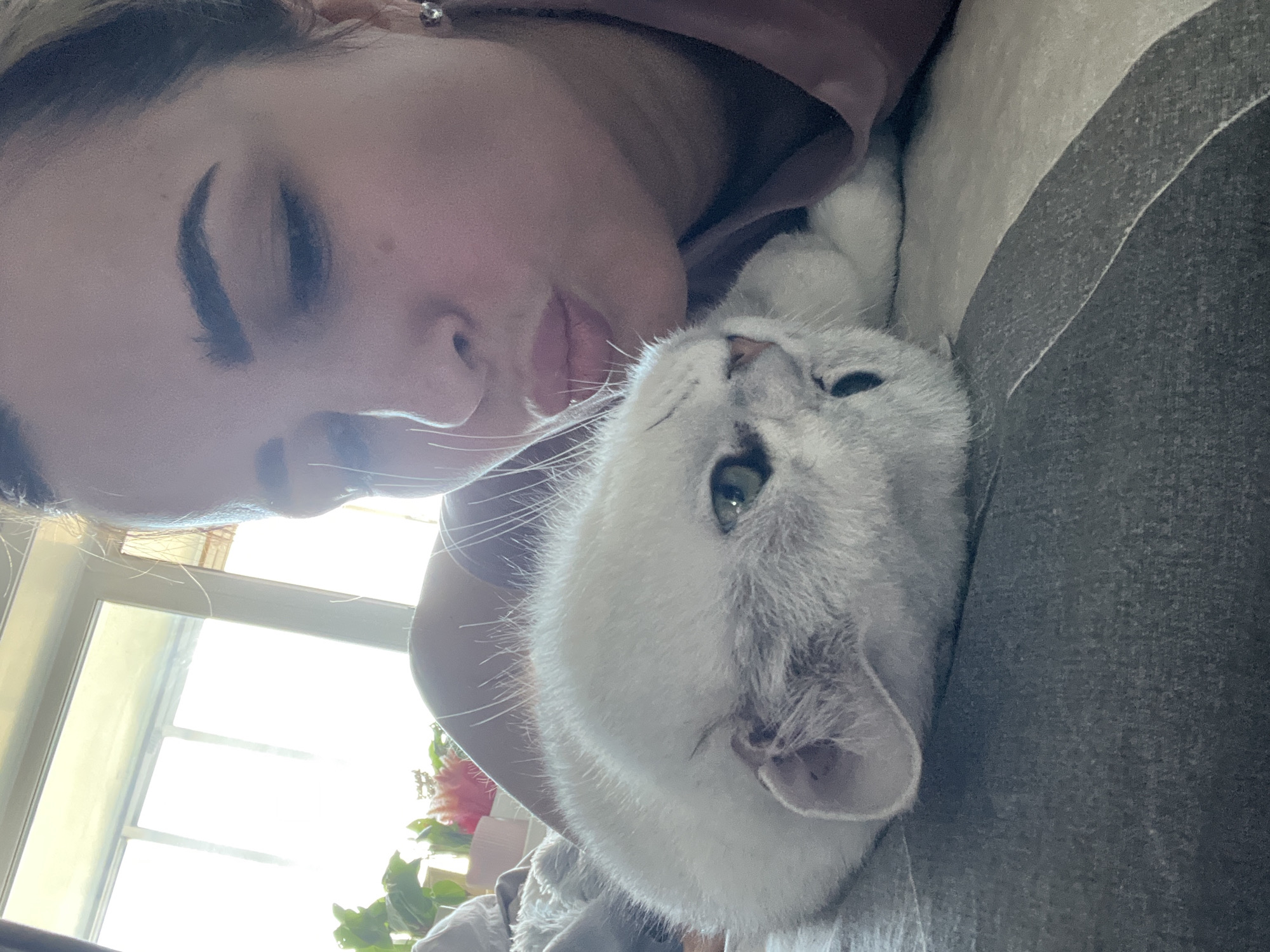 A selfie captures a woman and a white cat lying down. The woman's face is prominent, with her eyes closed and her lips near the cat. The cat, with its head tilted, has green eyes and a white coat. A window is visible in the background, and a plant can be seen near the window. The scene is indoors.