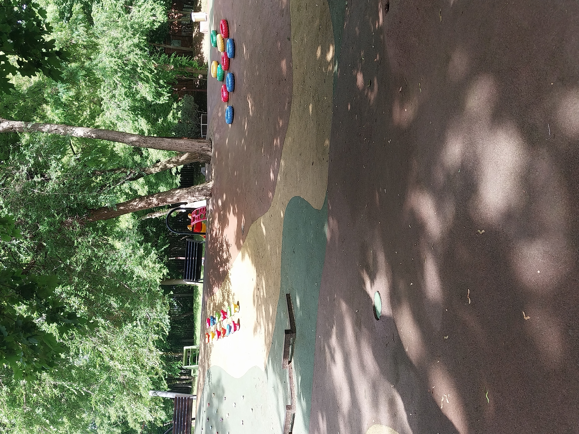 The image depicts an outdoor playground area with a soft, multi-colored surface. In the foreground, there are large, circular shadows cast by trees. The midground features a series of colorful stepping stones or small play elements arranged in a line, as well as a cluster of colorful rubber rings scattered on the ground. In the background, several park benches are visible, along with trees providing ample shade. The overall environment suggests a park or recreational area within the city of Dnipro, Ukraine, likely a children's play zone given the equipment. No individuals are present in the image.