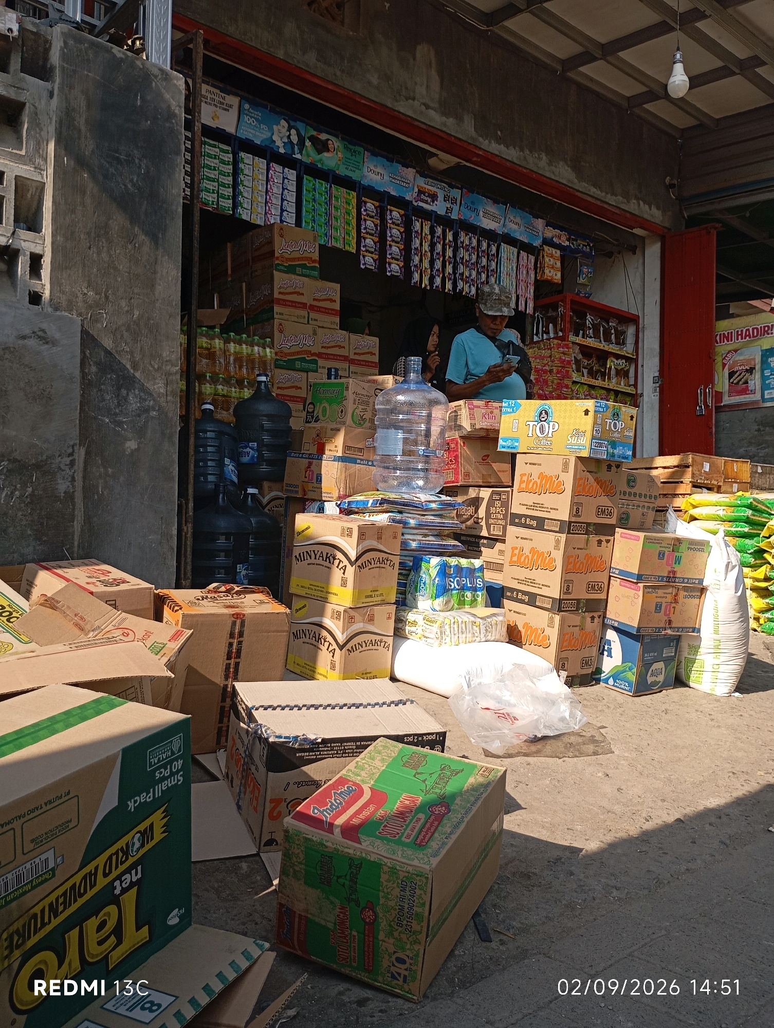 A street-level view of a small shop in Rambipuji, Indonesia, showcases a variety of packaged goods, both inside and stacked outside the store. The storefront features shelves lined with products such as bottled water, instant noodles, and cleaning supplies. In front of the shop, stacks of cardboard boxes and large plastic water containers are visible. A man wearing a blue shirt and a cap is standing inside the shop, looking at his phone. A woman in a black hijab is partially visible behind him. The ground is paved, and sunlight casts shadows, indicating it is daytime. The overall scene suggests a bustling local market or convenience store.