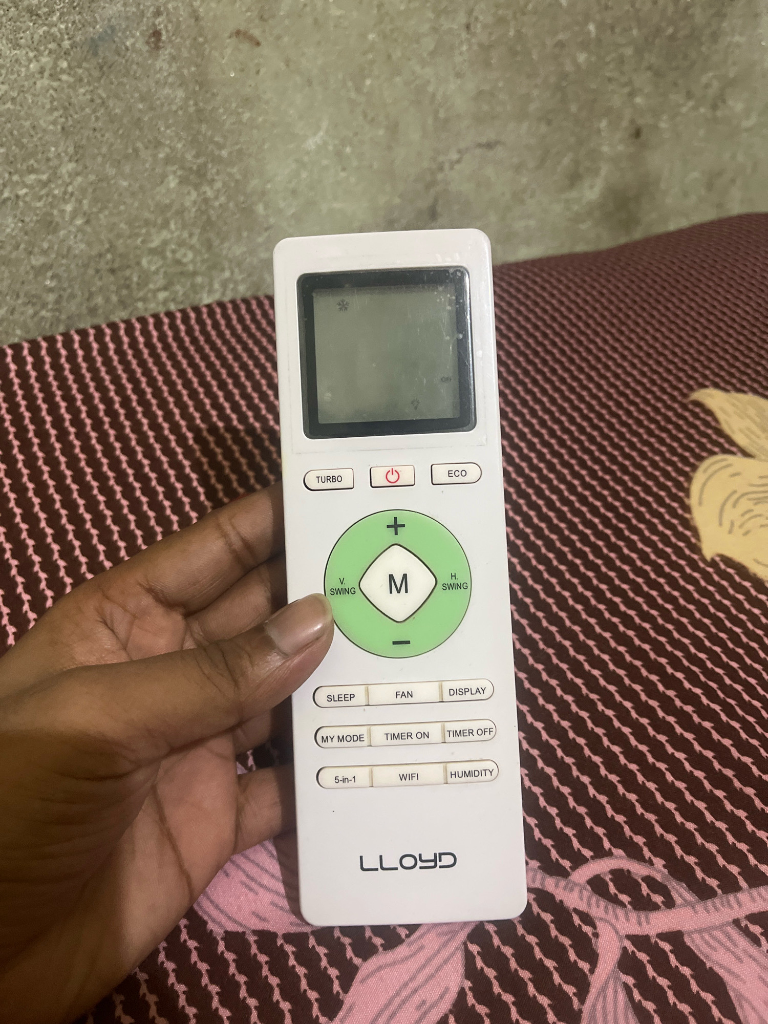 A hand is holding a white remote control for an air conditioner. The remote has a digital display screen at the top, followed by several buttons including "TURBO", "ECO", and a power button. Below these is a circular directional pad with "V. SWING" and "H. SWING" labeled on the left and right respectively, and a central button with "M" inside. Further down are buttons for "SLEEP", "FAN", "DISPLAY", "MY MODE", "TIMER ON", "TIMER OFF", "5-in-1", "WIFI", and "HUMIDITY". The brand name "LLOYD" is visible at the bottom of the remote. The remote is resting on a brown and pink patterned fabric surface, possibly a bedspread. The background is a textured, greyish-green wall. The overall scene appears to be an indoor environment. No specific location in Hisua, India, is identifiable from the image.
