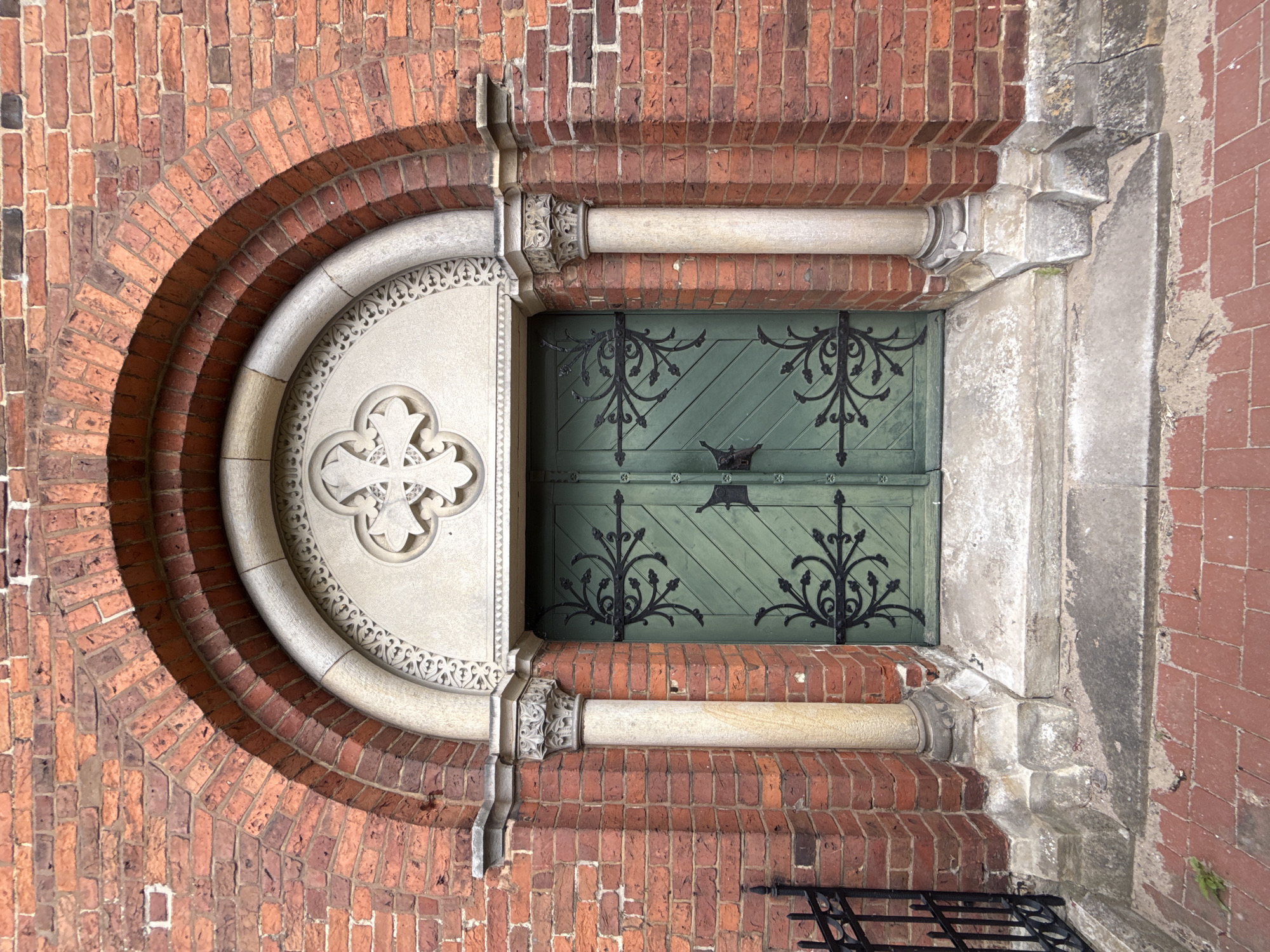 This is a close-up, eye-level, top-down shot of an ornate, historic doorway, likely part of a church or similar substantial building, located in Bassum, Germany. The primary materials visible are red brick and a light-colored, carved stone. The doorway itself is composed of two green wooden doors, featuring intricate black wrought-iron decorative elements and hinges. Above the doors, a large, round stone carving with a cross motif is set within a lighter stone surround, which is itself framed by a series of concentric brick arches. The brickwork shows some signs of age and weathering. The stone elements display detailed carvings and appear to be of a classical or gothic architectural style. The scene is well-lit, suggesting it was captured during the daytime, with no obvious indicators of specific weather conditions. The focus is entirely on the architectural details and the craftsmanship of the entrance, creating a sense of historical significance and artistry. There is no text visible in the image.