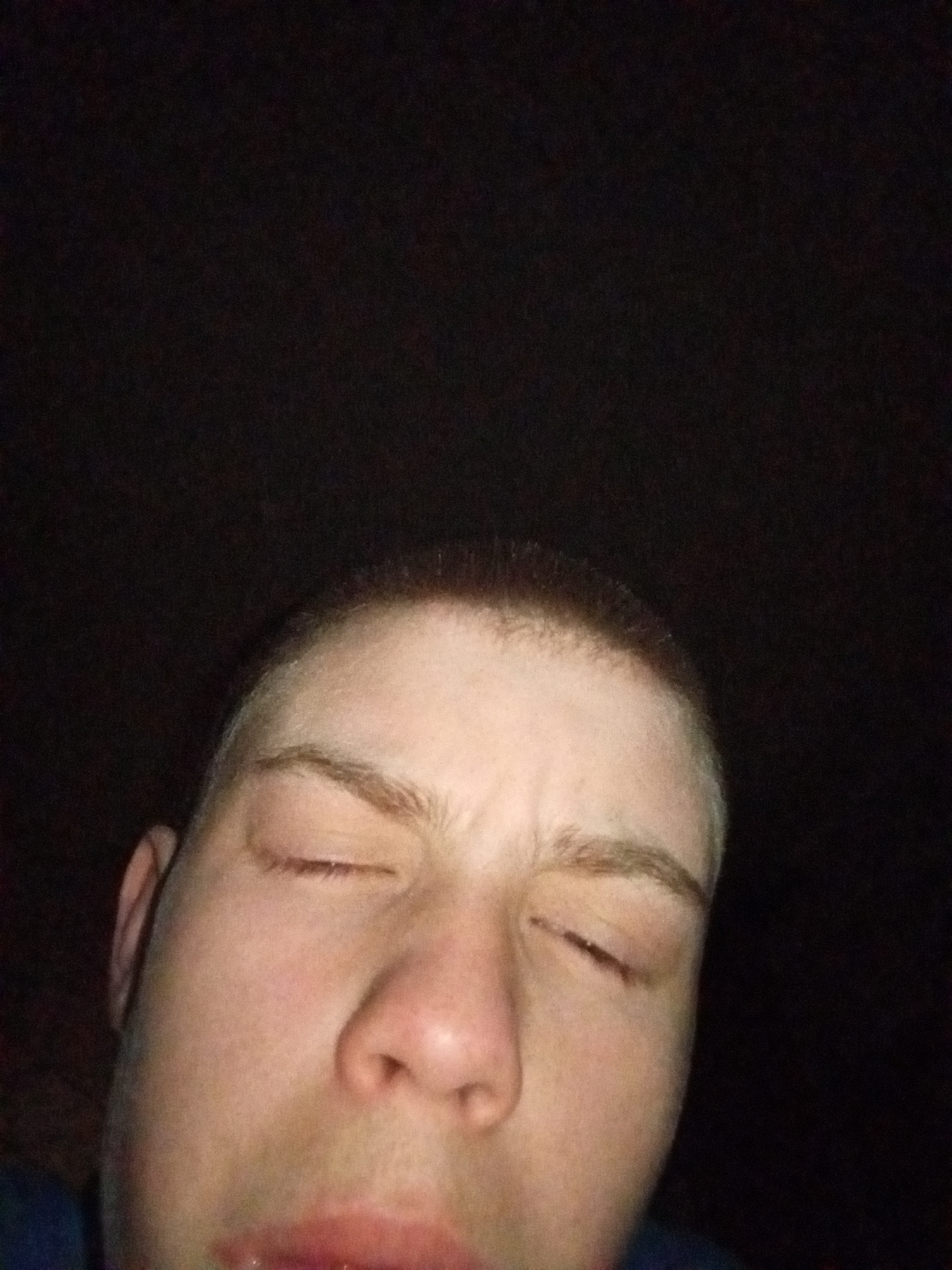 A close-up, poorly lit selfie of a fair-skinned person with their eyes closed. The person has short hair. The background is dark and undiscernible. There is no indication of the location being Щучье Озеро, Russia, or any other specific place.