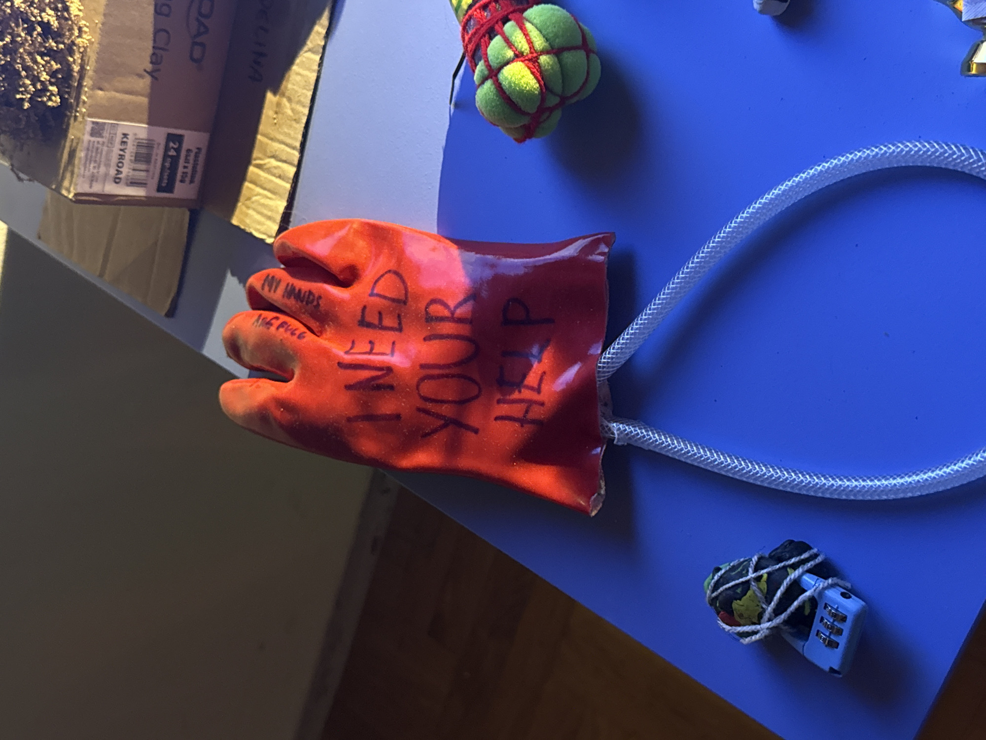 An overhead view shows a bright orange rubber glove on a blue surface. The glove is filled with a substance and has a clear plastic tube attached to its opening, leading away to the right. Handwritten black text on the glove reads, "I NEED YOUR HELP". To the right of the glove, a small, round object wrapped in red string is visible. Further to the right, a blue padlock secures a small, dark object wrapped in green and black material. In the background, a cardboard box with labels is partially visible, resting on a wooden surface. The lighting suggests an indoor setting with sunlight casting shadows.