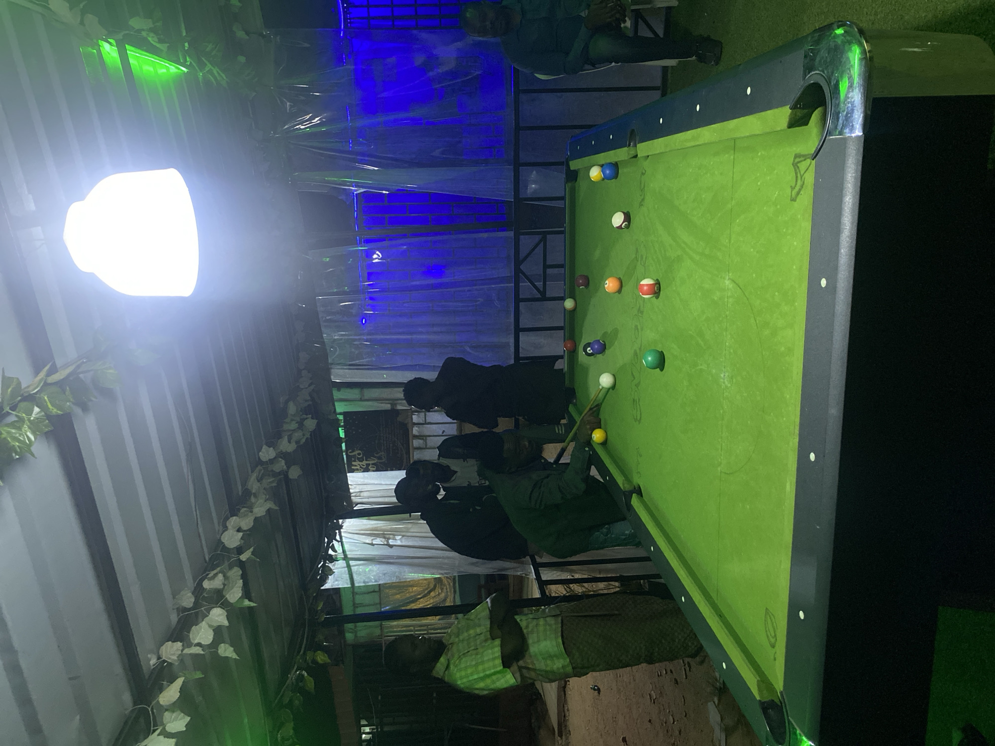 The image captures a nighttime scene at a casual outdoor or semi-outdoor venue, likely a bar or lounge, in Jos, Nigeria, where several men are engaged in a game of pool. The focal point is a green-felt pool table, which occupies the central and right portions of the frame, with various colored solid and striped balls scattered across its surface, along with a cue ball.
One man, wearing a green long-sleeved shirt, is actively playing pool, poised with a cue stick to take a shot. Two other men stand behind him, observing the game. In the left foreground, another man in a light plaid shirt stands, also watching the activity. A fifth individual is partially visible in the upper right background.
The setting is illuminated by a very bright, almost glowing, white light fixture positioned prominently on the left, casting strong light onto the scene. Complementary blue and green accent lighting is visible in the background, likely from LED strips, creating a vibrant, atmospheric glow. The venue features corrugated metal walls adorned with artificial ivy vines, giving it a rustic yet decorated feel. Translucent plastic sheeting or partitions are also visible in the background, suggesting a covered or partially enclosed space. The ground beneath the participants appears to be dusty earth, typical of an informal outdoor setting. The green felt of the pool table shows some signs of wear. The overall mood is social and relaxed. No discernible text is visible in the image.