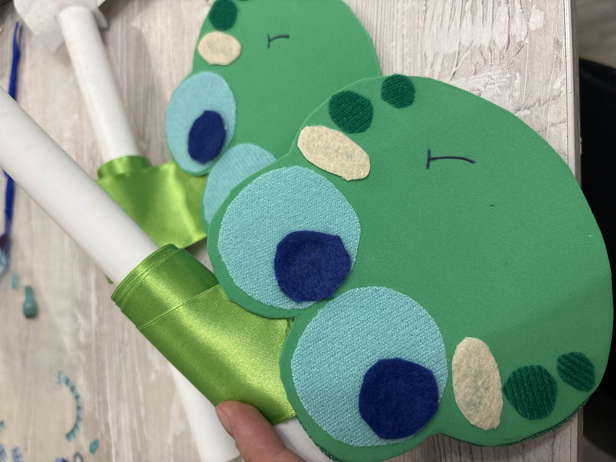 The image displays a close-up of two handmade frog faces, likely made of felt or foam, positioned on a light-colored wooden surface. The frogs are primarily green, with lighter green textured circles for their eyes, and smaller dark blue circles representing pupils. Some of the frogs have additional beige oval shapes and smaller dark green circles scattered on their faces, possibly representing cheeks or spots. One frog has a simple dark line drawn for a mouth.
In the background, two white cylindrical objects, possibly candles or rolls of material, are visible. One of these is partially wrapped with a shiny green ribbon, tied in a bow. The surface they rest on appears to be a wooden table with a textured, plank-like pattern. The overall lighting suggests an indoor setting, with the focus on the crafting materials.
Without additional information or context, it is impossible to determine if this image is located in the city of Izhevsk, Russia, or any other specific location.
