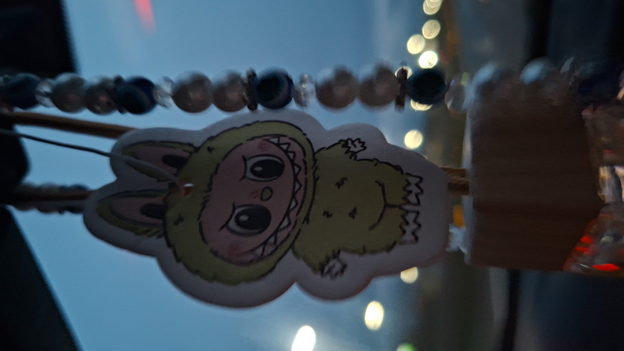 The image shows a fuzzy, monster-like sticker hanging from a beaded string. The sticker is predominantly green and cream-colored, with cartoonish features and large eyes. The beaded string consists of pearl-like white beads interspersed with dark blue beads. The background is blurred, with hints of city lights and a dusky blue sky. The photo was taken in a car. The sticker is hung in a car. It is difficult to provide an exact location within Самахвалавіцкі сельскі Савет, Belarus, as the image does not contain any distinctive landmarks or visual cues specific to a location within the village.