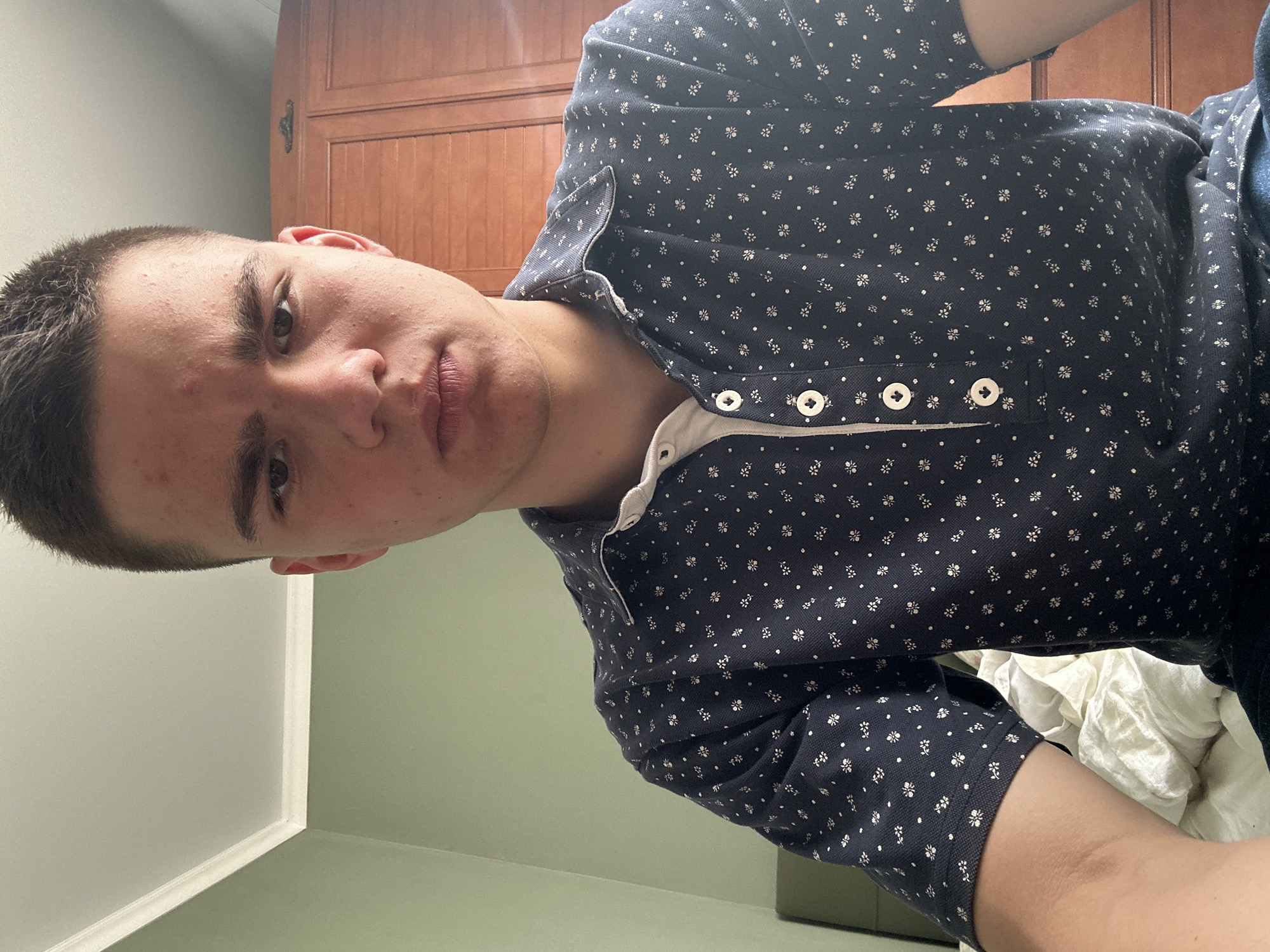 A close-up portrait shows a fair-skinned young man with a short, dark haircut. He is wearing a dark blue, short-sleeved shirt with a white floral pattern and a white placket with four visible white buttons. The background includes a light green wall, a white corner trim, a wooden cabinet, and crumpled white fabric, possibly a bedsheet. The location is an interior setting, city and location unknown.