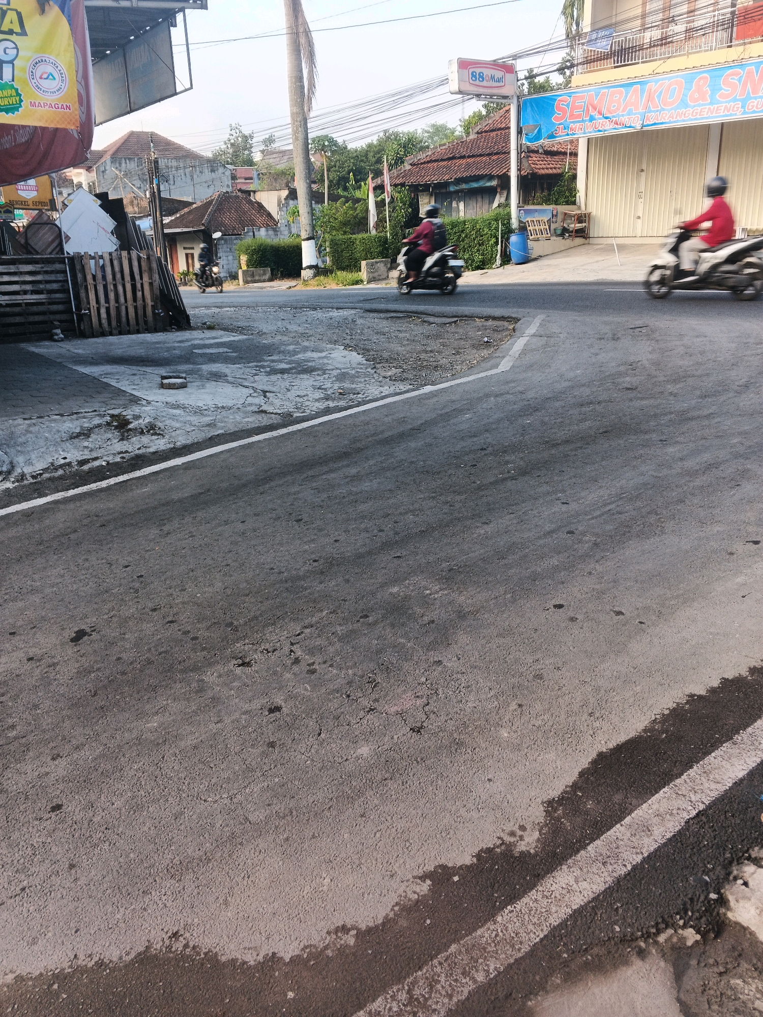 In Semarang, Indonesia, this outdoor scene captures a street with two individuals on motorcycles. One motorcycle, ridden by a person in a red shirt, is blurred in motion on the right side of the road. To the left, another motorcycle, ridden by a person in a dark outfit, is also blurred, moving in the same direction. In the background, a building with a large blue banner reading "SEMBAKO & SN" is visible, along with a 7-Eleven convenience store sign. Wooden pallets are stacked on the left side of the image. The road surface appears worn, with some patchy areas and a white line marking the lane. The environment suggests a commercial area within the city.