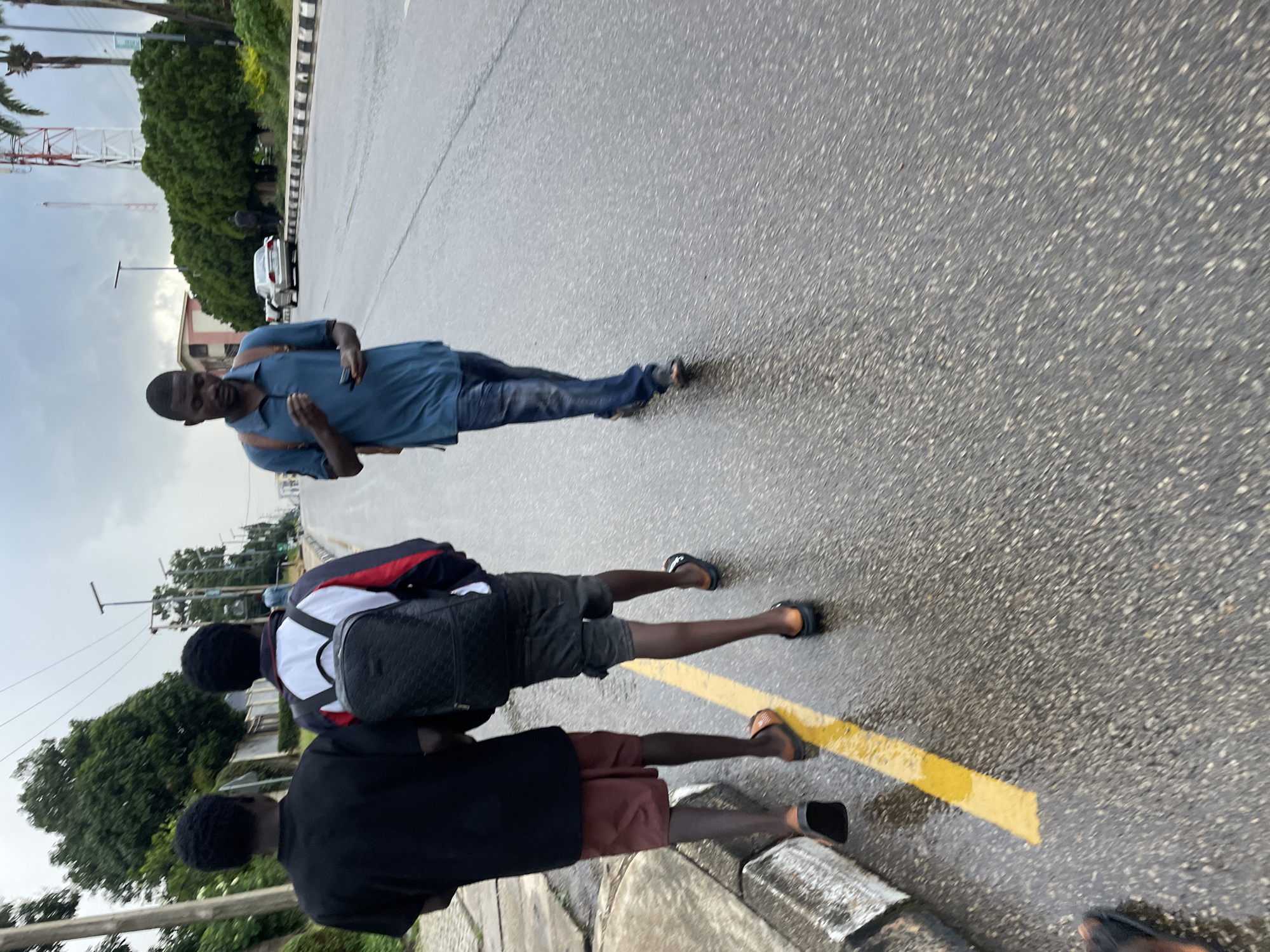 In Jos, Nigeria, three young men are walking down a wet asphalt road, indicating recent rainfall. Two of the men walk side-by-side, closer to the sidewalk which features a yellow painted line. The man on the left wears a black t-shirt and red shorts, while the man on the right wears a jacket with white, red, and blue accents over a white shirt, and grey shorts. He carries a black backpack with a quilted pattern. The third man walks ahead of them, slightly to the right, in the middle of the road. He is wearing a blue polo shirt and blue jeans, and is holding a mobile phone in his right hand. A light-colored sedan is visible in the background, further down the road. The environment appears to be a suburban or semi-urban area, with trees and buildings lining the street, and a communication tower visible in the distance. The sky is overcast, suggesting a cloudy or rainy day.