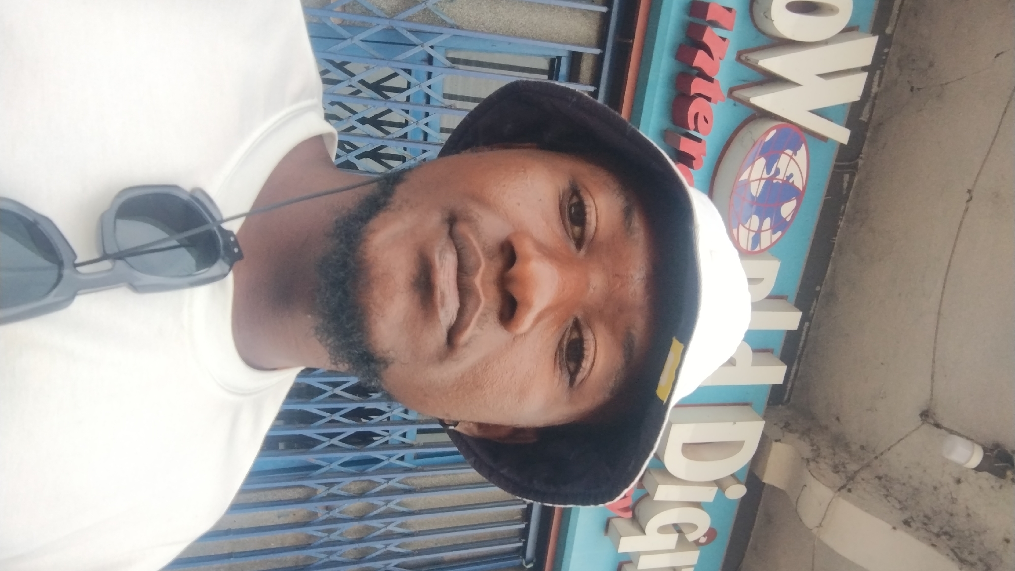 A man with dark skin, a short black beard, and wearing a white T-shirt and a white bucket hat with a dark brim, is photographed outdoors in Jos, Nigeria. Sunglasses are hanging off the shirt. The background includes a blue metal gate and a sign that reads "Interworld Digital" in white letters on a blue background, featuring a logo of the Earth. A bare lightbulb fixture is above the sign. The image is rotated clockwise.