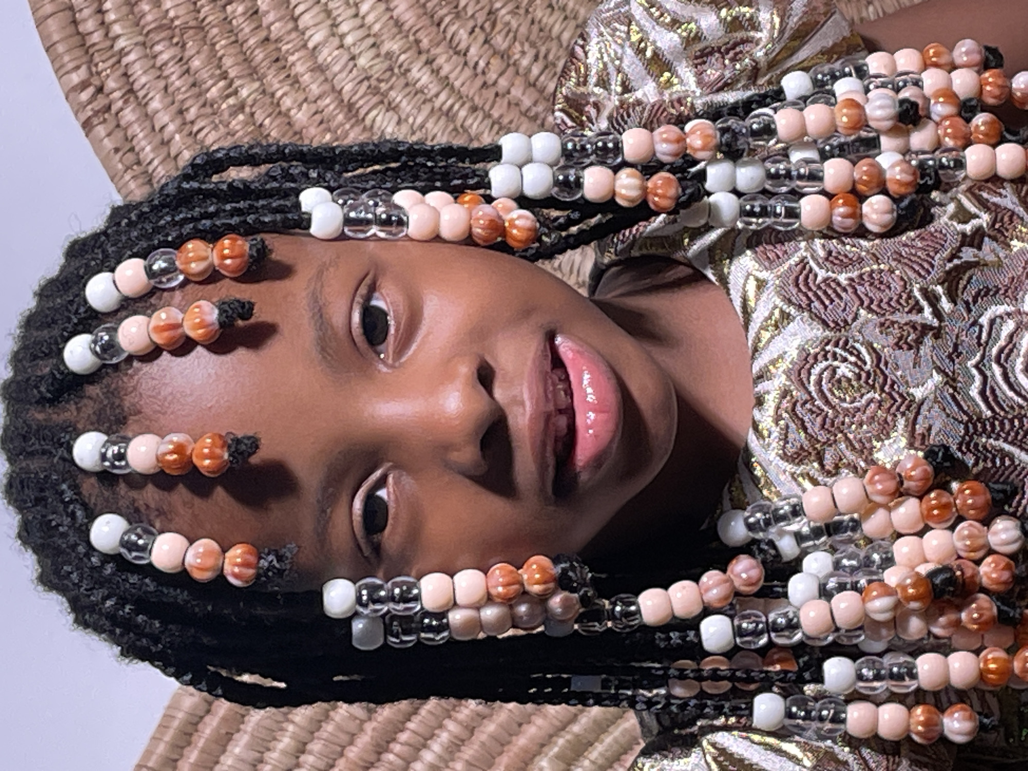 A young person with dark skin is lying down and looking directly at the camera. They have braided hair adorned with beads of various colors, including white, clear, peach, and orange. The individual is wearing a patterned gold and brown garment. They appear to be lying on a woven mat. There is no clear indication that this photo was taken in Barakin Akawo, Nigeria. The image provides no forensic context.