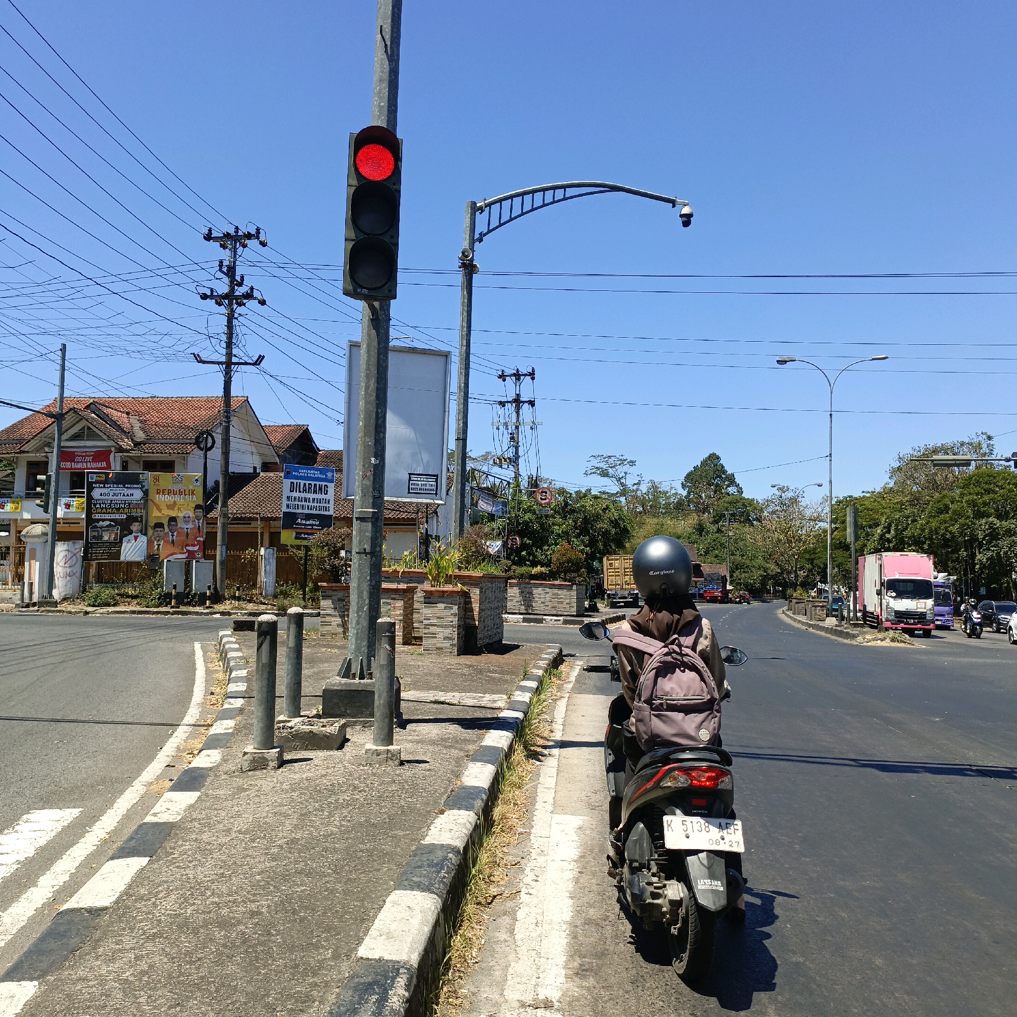 The image shows a street intersection in Salatiga, Indonesia, with a traffic light displaying red. A person on a scooter, wearing a helmet and a backpack, is stopped at the intersection, facing away from the camera. The scooter has a license plate that reads "K 5138 AEF". In the background, there are buildings, trees, power lines, and other vehicles on the road. A security camera is mounted on a pole above the intersection. The sky is clear and blue.