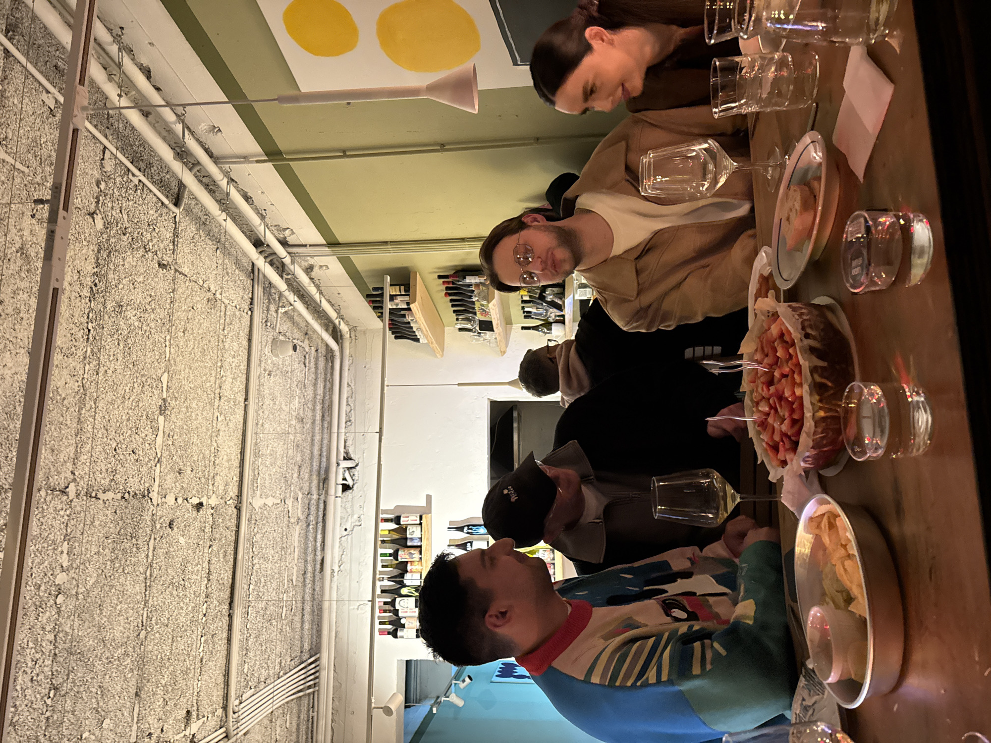A group of six people are seated at a long, narrow wooden table in what appears to be a restaurant or bar in Belgrade, Serbia. The scene is indoor and lit with a mix of ambient and warmer artificial light. The walls are a mix of exposed concrete, white plaster, and painted sections of light blue and green with yellow circles painted on the green section. Wine bottles are lined up on shelves along the walls.
On the table, there are wine glasses, water glasses, plates with food, and a serving bowl filled with a red fruit, possibly strawberries. The people are dressed in casual clothing, some wearing jackets and hats. They appear to be engaged in conversation and eating, enjoying a meal together. One person is wearing glasses and a tan jacket. The location is identifiable as Belgrade due to architectural styles and elements, possibly a trendy or modern establishment.