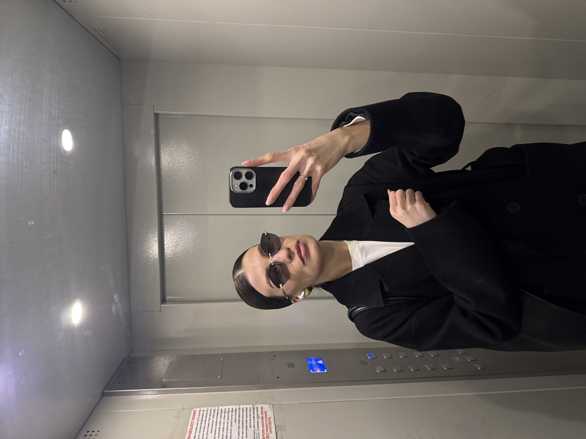 The image is a selfie taken in an elevator. A person with slicked-back hair and sunglasses is holding a black smartphone up to take the photo. They are wearing a black coat over a white shirt. The elevator has grey walls and control buttons. There is a sign with text in Russian. The location appears to be in the city of Chaikovsky, Russia, based on the presence of Russian text.