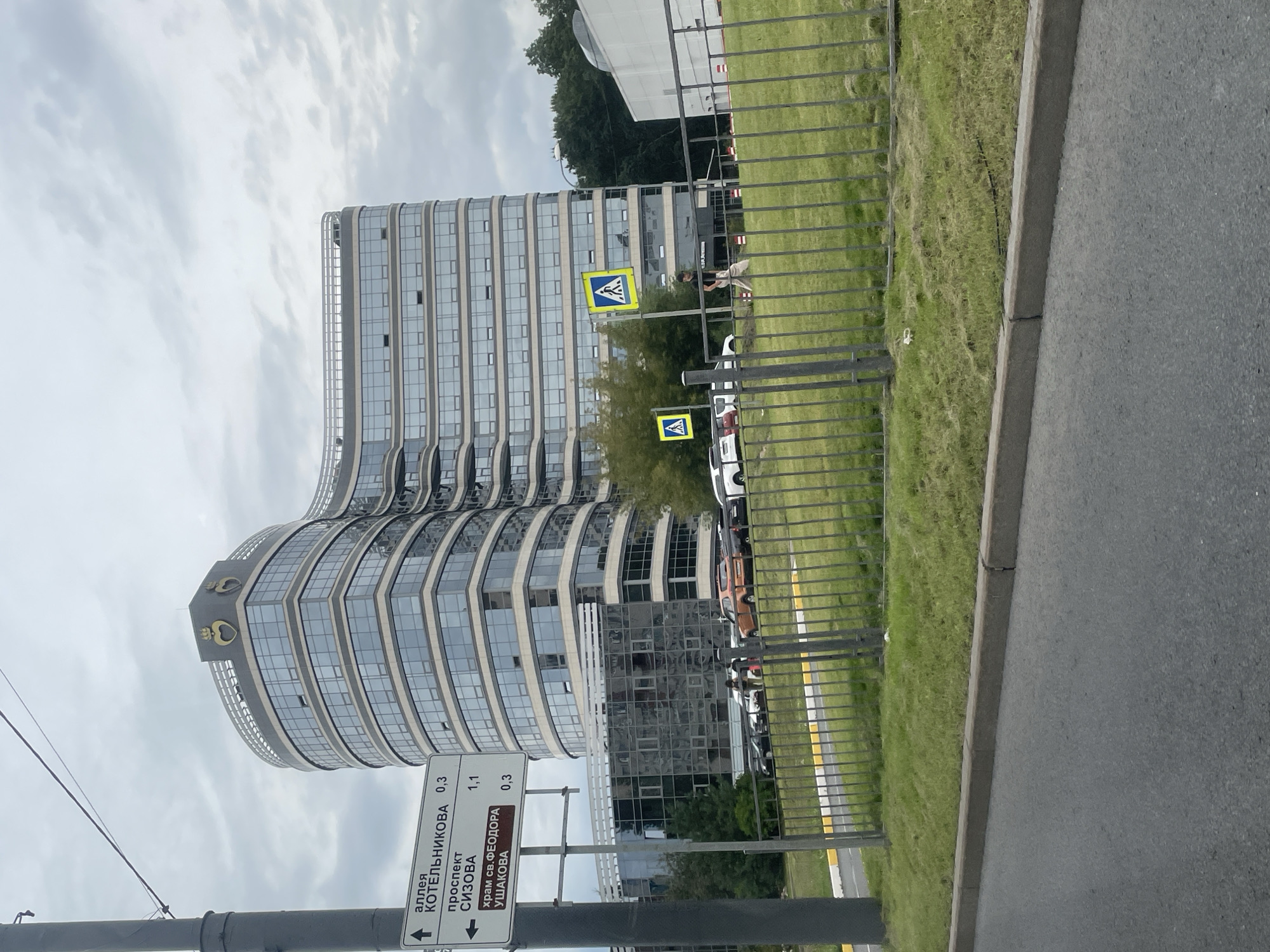 A modern, multi-story building with a curved facade and numerous windows is the central focus of the image, located in the city of Saint Petersburg, Russia. The building has a rounded top with a decorative element resembling a crown and a heart. In the foreground, a street with a grey asphalt surface and a concrete curb is visible. A metal fence runs along the edge of a grassy area. A traffic signpost with directional arrows and street names in Russian stands to the left of the building. Several cars are parked near the building. Two pedestrian crossing signs with yellow backgrounds and white pedestrian symbols are also present. A person in dark clothing is walking across the street, and another person can be seen in the distance near the parked cars. The sky is overcast with clouds.