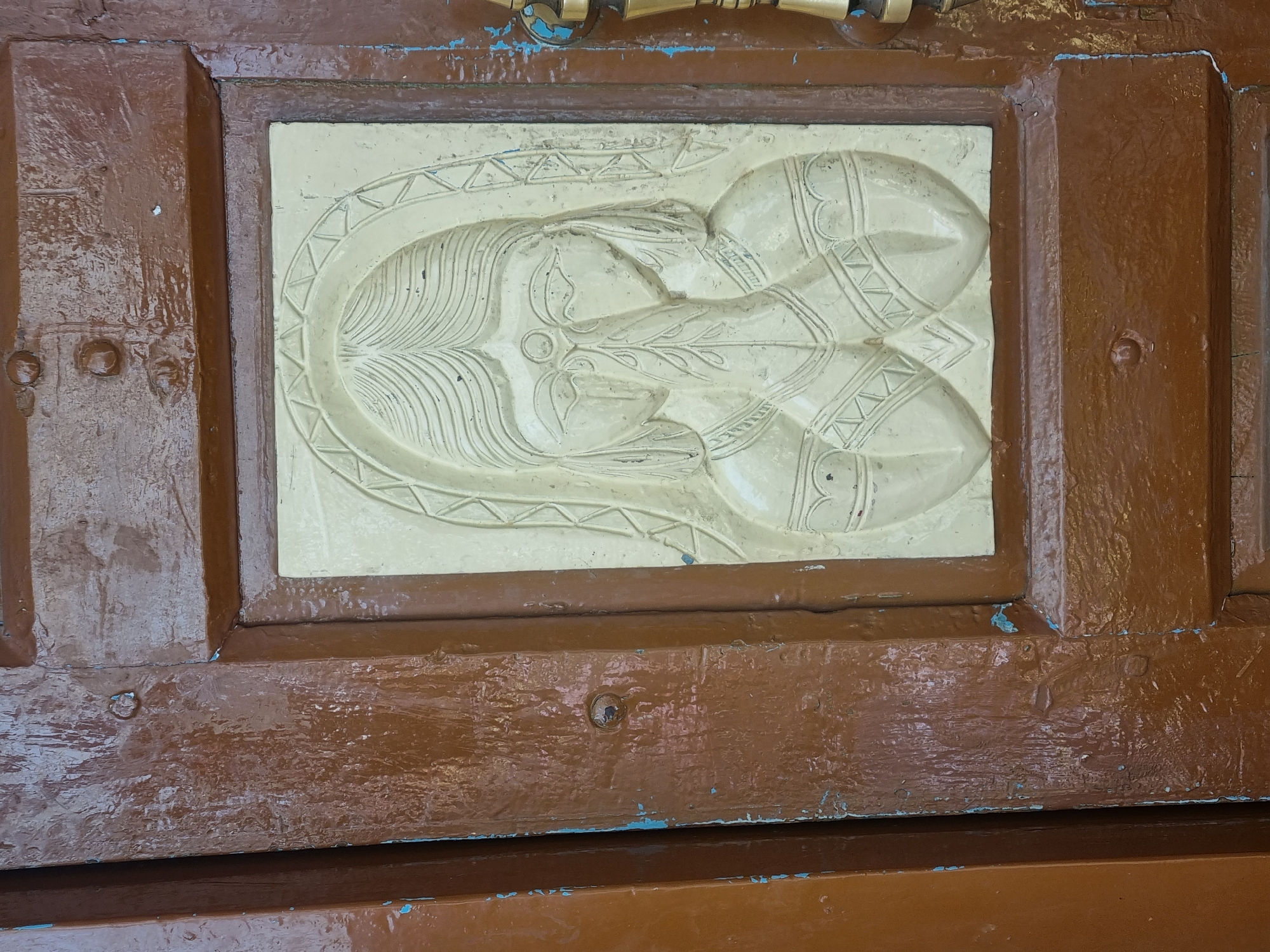 Located in Pedda Kammavaripalli, India, the scene displays a close-up of an aged brown wooden door. Embedded centrally within a paneled section of the door is a rectangular, light beige bas-relief carving measuring approximately 30-40cm in height. This carving depicts a stylized female figure from the bust upwards, facing forward. The figure features long, flowing hair and an ornate, arching headpiece or veil. Her hands are pressed together in front of her face, concealing her mouth and chin, in a gesture suggestive of prayer or greeting. Decorative patterns are visible on her attire and arms, resembling jewelry or clothing embellishments. The surrounding wooden door exhibits significant wear, with chipped and peeling brown paint, revealing underlying traces of a lighter blue pigment in multiple areas. Small, dark circular indentations or protrusions are visible on the door's wooden frame, particularly in the upper and lower left quadrants. A gold-toned metallic door handle or latch mechanism is partially visible along the right edge of the door.