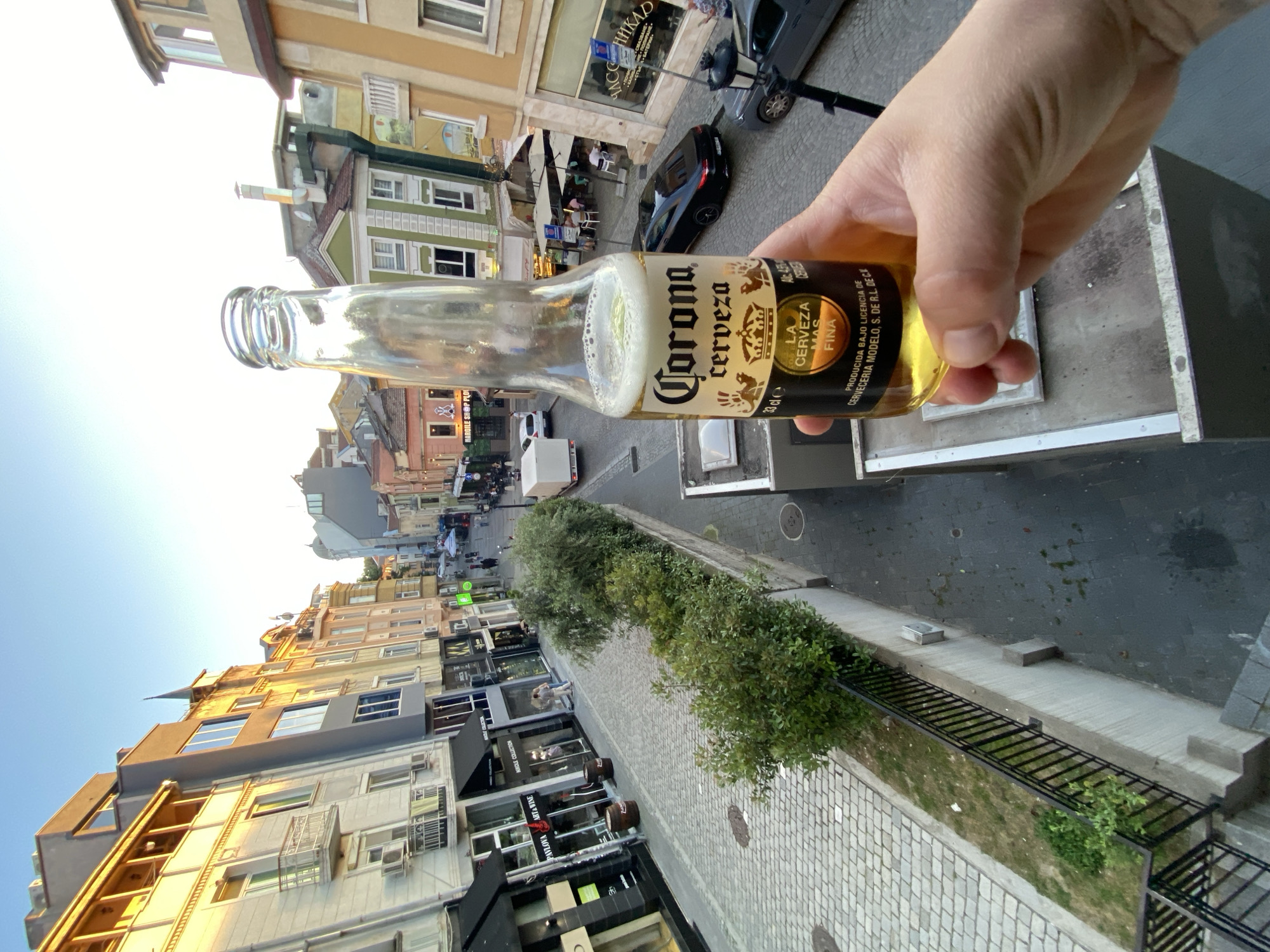 The image is taken from an elevated position, possibly a balcony, overlooking a street in the city of Plovdiv, Bulgaria. The central focus is a hand holding a partially consumed bottle of Corona beer. The street scene features a mix of architectural styles, with several multi-story buildings lining both sides. Businesses are visible at street level, including a "NIKOLE COLLECTION" store, "PAVLOVA ART & WINE", and "HARGILE SHOP PLOI." There is also a sign that reads "ЧАССНИКАD" «СТЪКА-БАТЕРИИ». A row of trimmed bushes separates the street from a pedestrian walkway paved with stones. Vehicles, including cars and a scooter, are parked on the street. Several people can be seen walking along the street. The sky is clear, suggesting fair weather conditions.