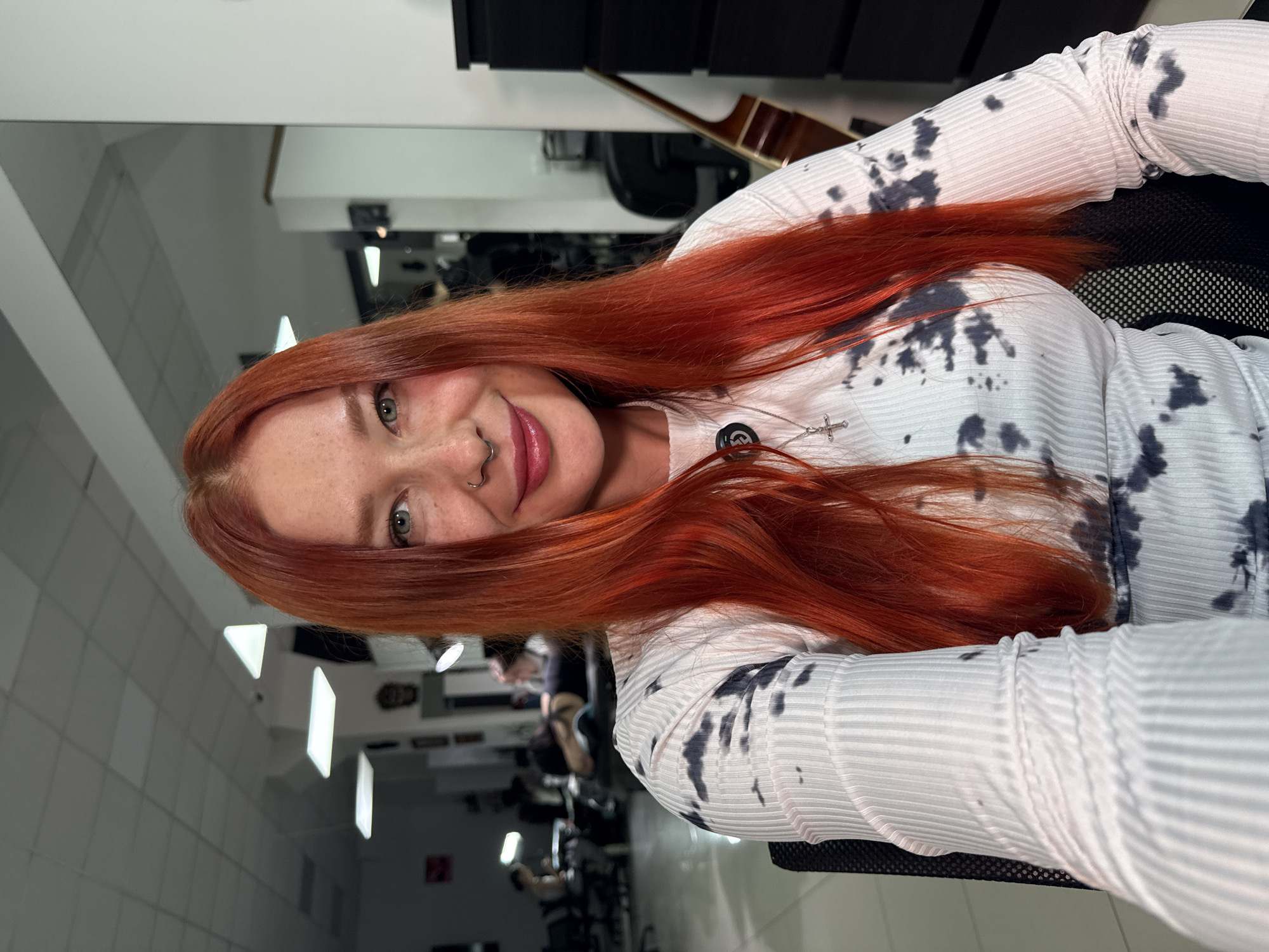 A selfie taken indoors in Rostov-on-Don, Russia, features a woman with red hair. She has fair skin with freckles, a pierced nose, and is wearing a long-sleeved white shirt with black tie-dye patterns. A necklace with a small cross pendant is visible. The background reveals a room with mirrored walls, reflecting other individuals and fitness equipment, suggesting it is a gym or fitness studio. A guitar is visible behind her head. The room is brightly lit by overhead fluorescent lights.
