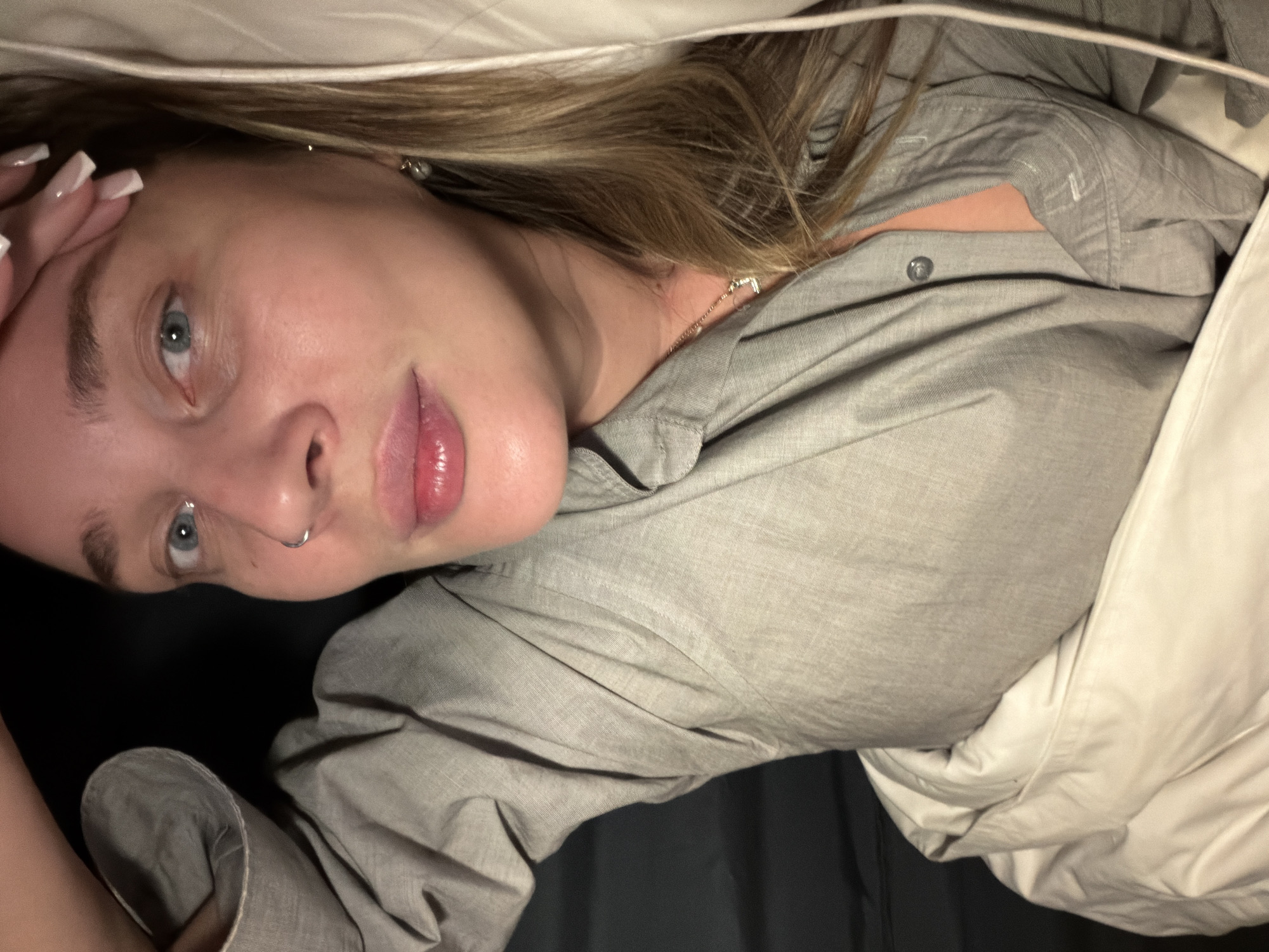 The image presents a portrait of a young adult female, likely located in Moscow, Russia, lying down and looking directly at the viewer. Her light blonde or light brown hair frames her face, and her eyes are a distinct blue. She wears a light grey, long-sleeved collared shirt. Her left hand is raised towards her head, showcasing long fingernails with white tips.
Notable details include a small silver septum piercing, a delicate silver necklace with a tiny pendant visible on her chest, and small silver earrings. She appears to be in bed, partially covered by a white blanket or duvet that runs across her body and past her head. The background is dark, suggesting an indoor setting with minimal ambient light. Her facial expression is neutral and calm, with a direct gaze towards the camera. There is no visible text in the image.