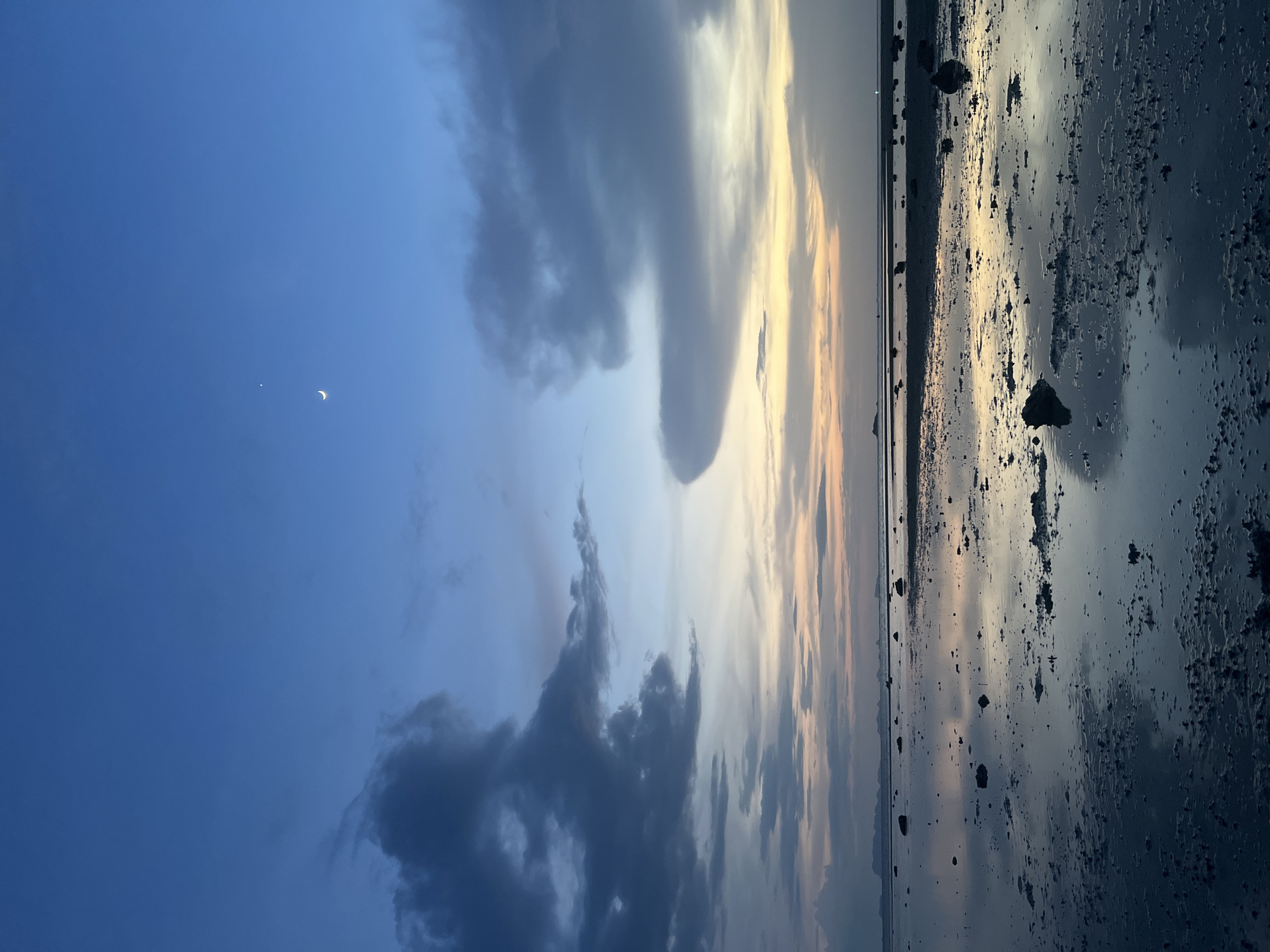 This image depicts a serene natural landscape during twilight, likely in the Ko Pha-ngan District of Thailand. The sky is a gradient of deep blues near the top, transitioning to softer blues and hues of orange and yellow near the horizon, suggesting the sun has recently set or is about to rise. A slender crescent moon is visible high in the sky, accompanied by a single bright star. Dark, heavy clouds are scattered across the sky, adding a dramatic element to the scene.
The foreground is a shallow body of water, possibly the sea at low tide, reflecting the colors of the sky. The water's surface is not perfectly smooth, with small ripples and textures on the muddy or sandy bottom. Scattered across this reflective surface are numerous dark, irregular-shaped rocks of varying sizes. In the distance, silhouetted against the horizon, are the faint outlines of islands or landmasses. The overall impression is one of quiet solitude and the beauty of nature at dusk. There are no people or animals visible in the frame.