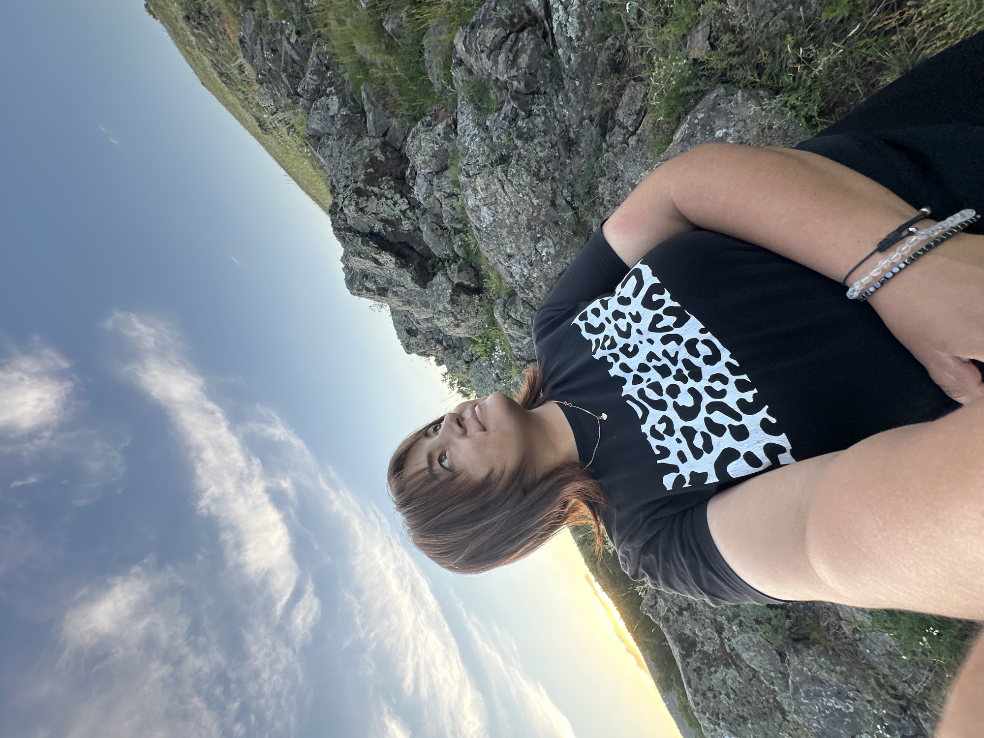 A woman is lying on her side, looking up at the sky. She is wearing a black t-shirt with a white leopard print design on it, and several beaded bracelets on her wrist. She is on a rocky surface, with grass and a cliff face behind her. The sky is blue with some wispy white clouds, and the sun appears to be setting, casting a warm glow on the horizon. The overall mood is peaceful and contemplative.