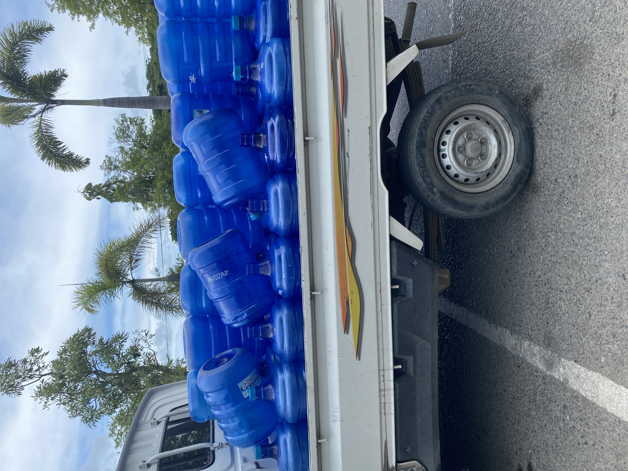The image shows a white pickup truck loaded with many blue plastic water jugs, parked on a paved road. The jugs are stacked several layers high and fill the truck bed. Some of the jugs have labels visible. In the background, there are trees, including palm trees, and a cloudy sky. The scene appears to be outdoors in a tropical or subtropical location. The truck has a decal on its side with yellow and orange stripes. The overall impression is of a delivery or transport of bottled water in what is likely a city or town in Banda Aceh, Indonesia, given the vegetation and architecture typical of the region.