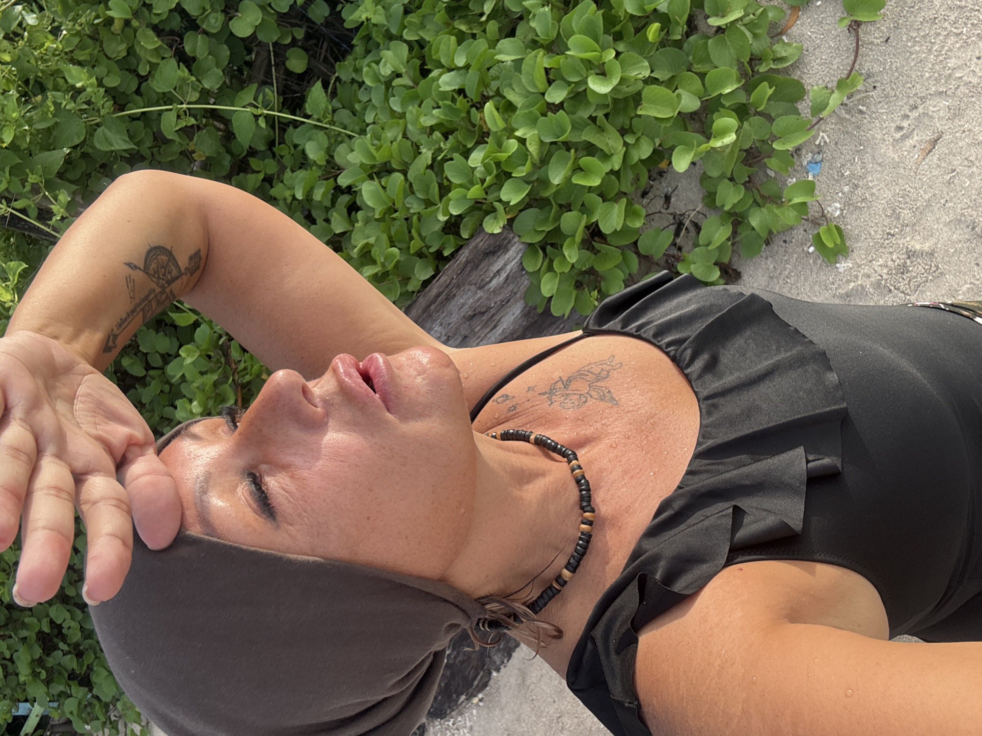 A woman lies on her back near vegetation and sand. She is wearing a dark, sleeveless top and a head covering. A beaded necklace adorns her neck. Tattoos are visible on her chest and arm. Her hand is near her forehead, possibly shielding her eyes. The location is likely near a beach or sandy area in Denpasar, Indonesia, indicated by the presence of sand and coastal vegetation.