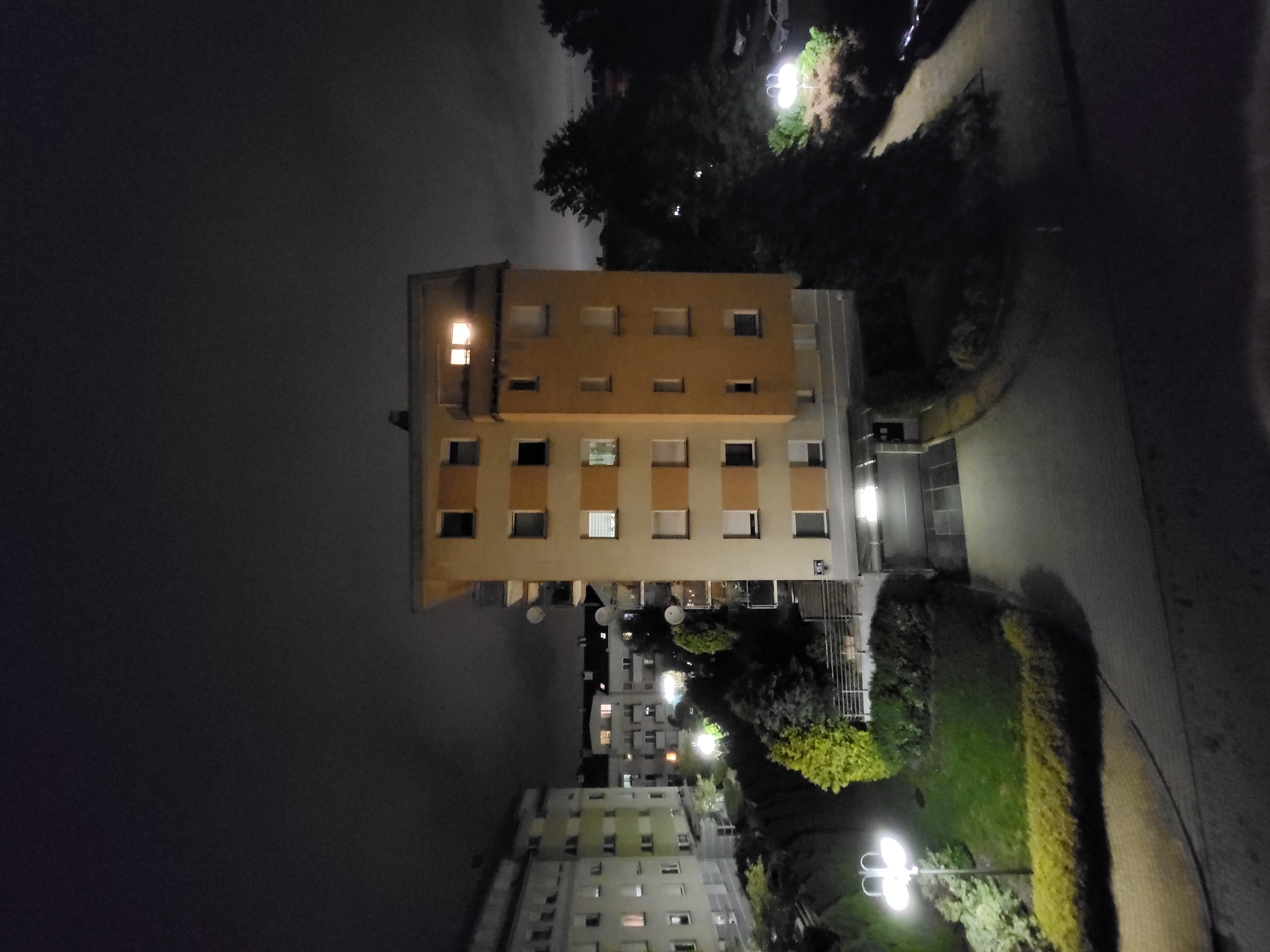 The image captures a nighttime scene of a residential area, likely an apartment complex, under a dark and possibly overcast sky. The photograph is oriented vertically, showing buildings and illuminated pathways.
In the foreground, a prominent multi-story building with a light brown or beige facade is visible. This building features multiple rectangular windows across its several floors; some windows are brightly lit from within, indicating occupancy, while others are dark or have blinds drawn. One window on the upper left side of this building is particularly luminous, casting a warm glow.
To the right of the main building, a paved pathway curves through the scene, flanked by green lawns and neatly trimmed hedges. Several streetlights, characterized by their dual-globe fixtures, brightly illuminate sections of the path and the surrounding vegetation, creating stark contrasts between light and shadow. Dark, dense trees are scattered throughout the landscape, contributing to the nighttime atmosphere.
In the background, additional multi-story residential buildings can be seen, also displaying illuminated windows and suggesting a broader urban or suburban setting. Some of these distant buildings appear to have satellite dishes attached to their exteriors. The overall impression is one of a quiet, illuminated residential environment after dark. No human activity or legible text is visible in the image.