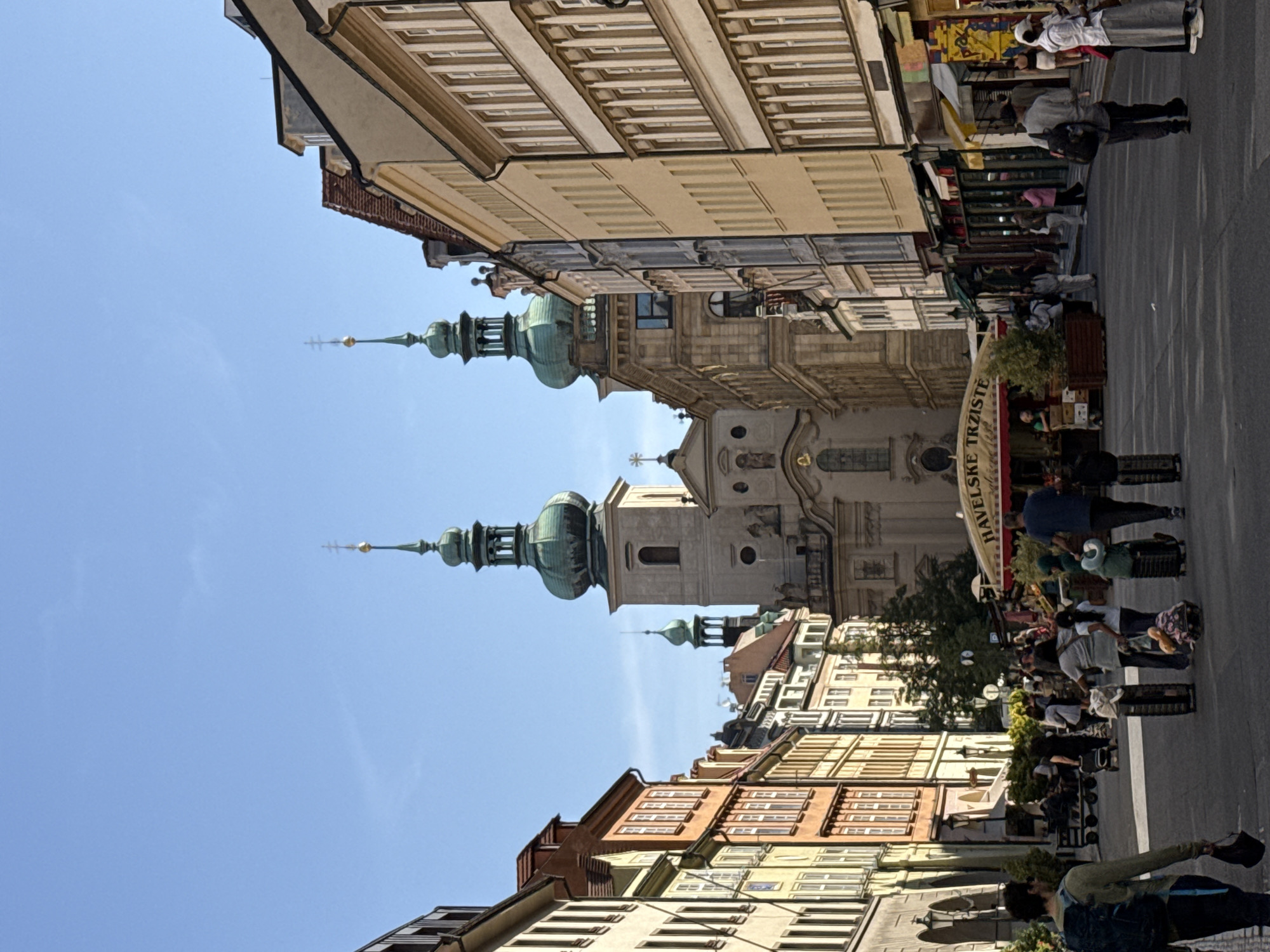 The image shows a street scene in Prague, Czechia, featuring the Church of Our Lady before Týn in the background. The church's twin Gothic spires dominate the skyline. The street is lined with colorful historic buildings, many of which appear to be shops with awnings and displays. A market stall with a yellow awning and the text "HAVELSKE TRZISTE" is visible in the midground. Several people are walking along the street, some with luggage, suggesting it is a tourist area. The sky is clear and blue.