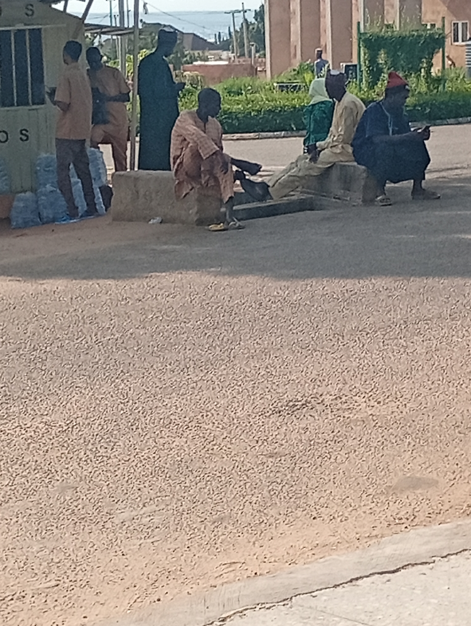 The image depicts a street scene in Gombe, Nigeria, with several individuals gathered. In the foreground, a person is seated on a rough, concrete structure, looking to their right. To their left, a group of four men are positioned. Two men are standing, one in a tan outfit and another in a darker brown outfit, seemingly interacting near a structure with signage that includes the letters "S" and "OS". Piles of blue plastic bags are visible next to them. Another man, dressed in a brown and white patterned garment, is seated on a similar concrete structure. Further to the right, two men are seated on a concrete ledge. One is wearing a light-colored robe and a cap, while the other is dressed in dark clothing and a red cap, holding a mobile phone. The background shows buildings, vegetation, and a slightly elevated horizon line, suggesting an urban or semi-urban setting. The ground is covered with a rough, textured surface.
